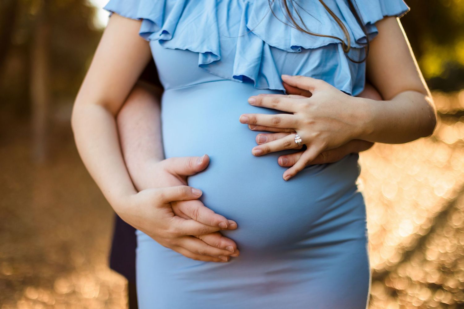 Hands gently rest on a pregnant belly wrapped in a light blue maternity dress during an outdoor sunset photoshoot.