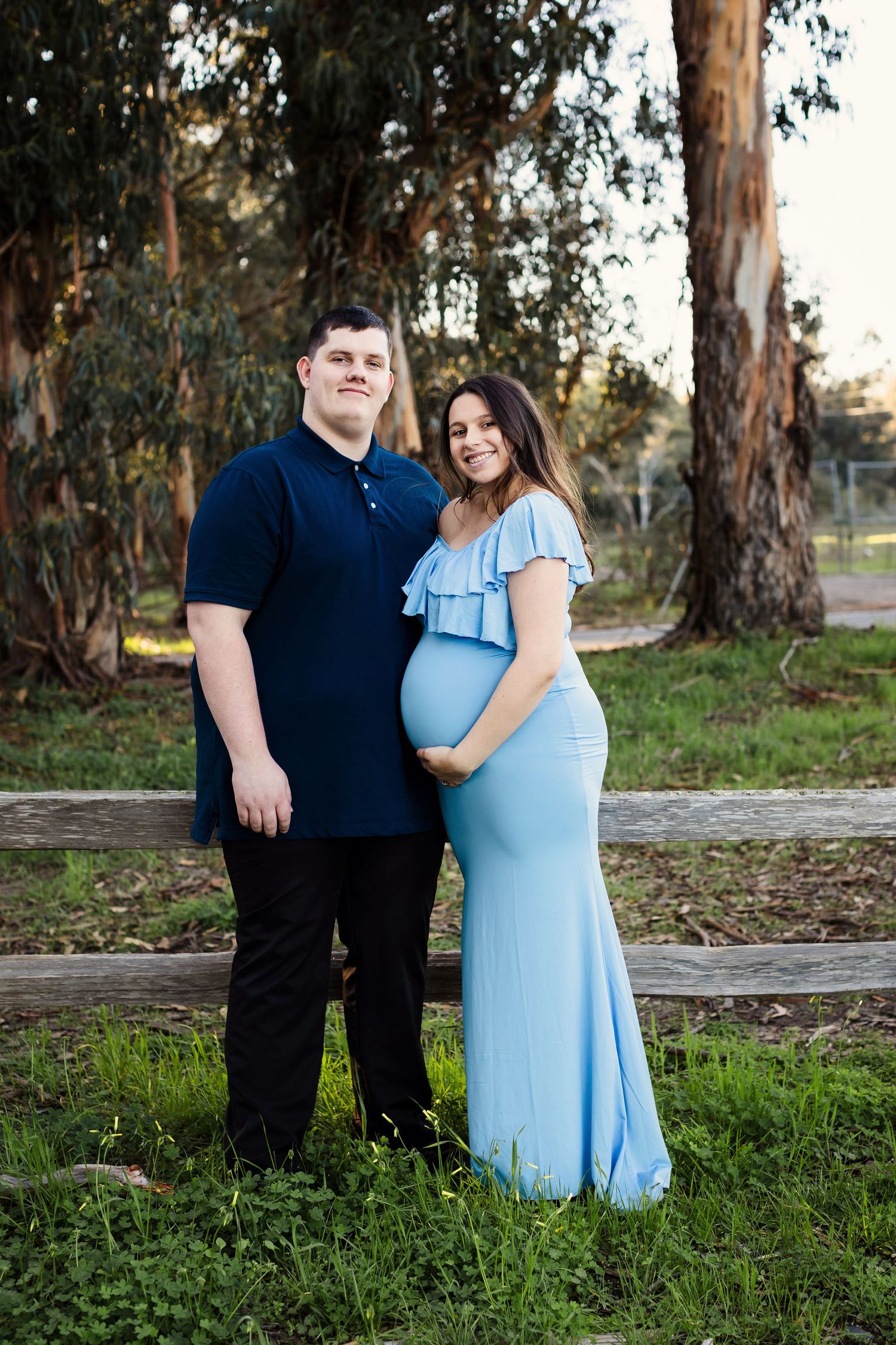 A couple stands together in casual attire against a backdrop of tall eucalyptus trees at sunset.