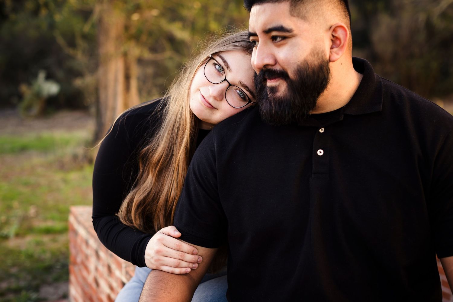 A couple embraces outdoors on a sunny day, with one wearing glasses and a black sweater leaning on their partner.