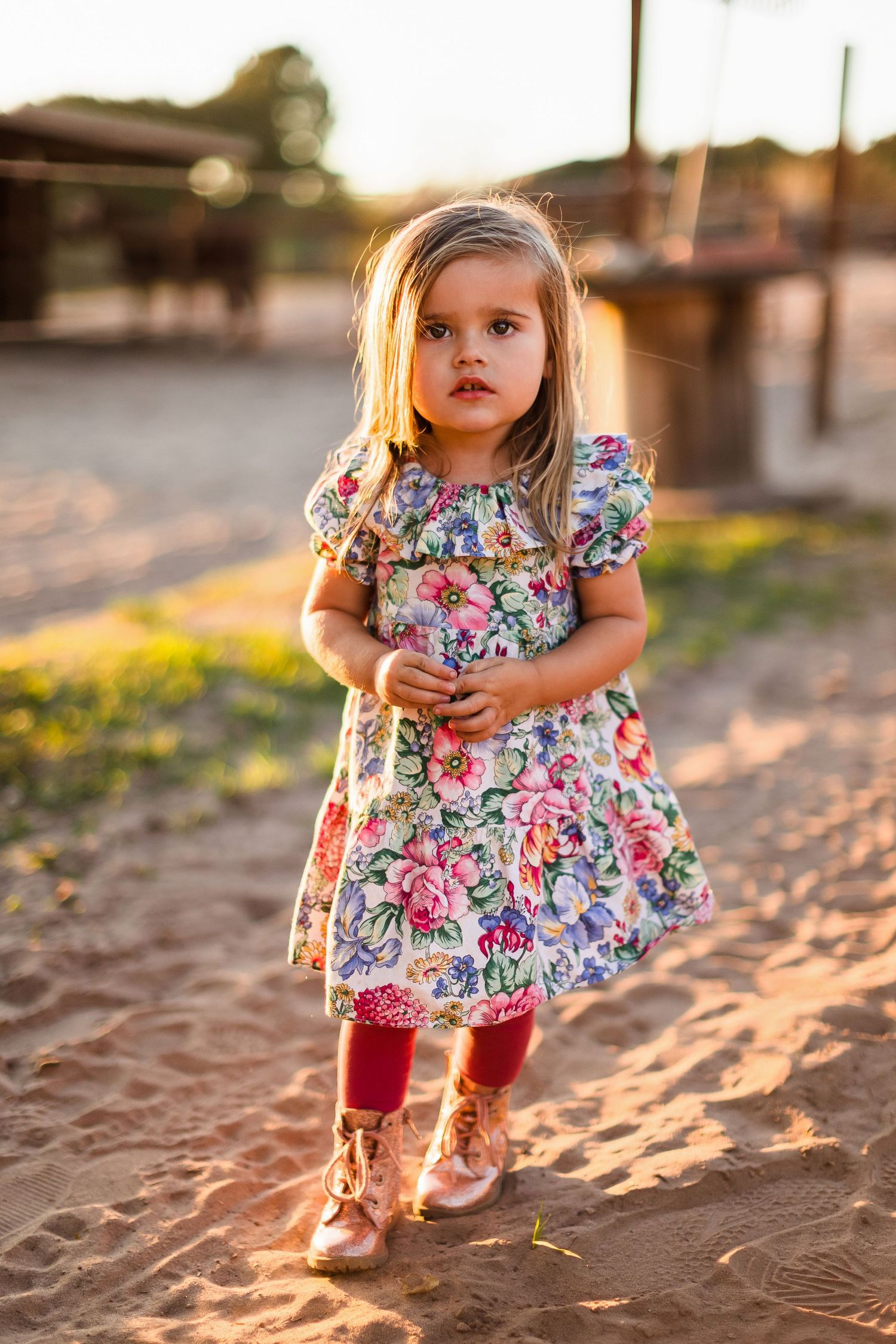 Child wearing floral dress plays in golden sunlight casting long shadows on sandy ground during sunset.