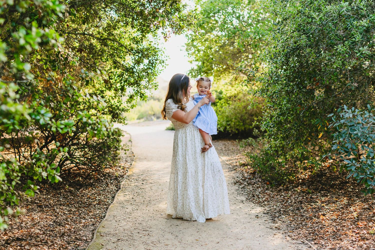 A couple walks together along a garden path surrounded by lush greenery and trees on a sunny day.