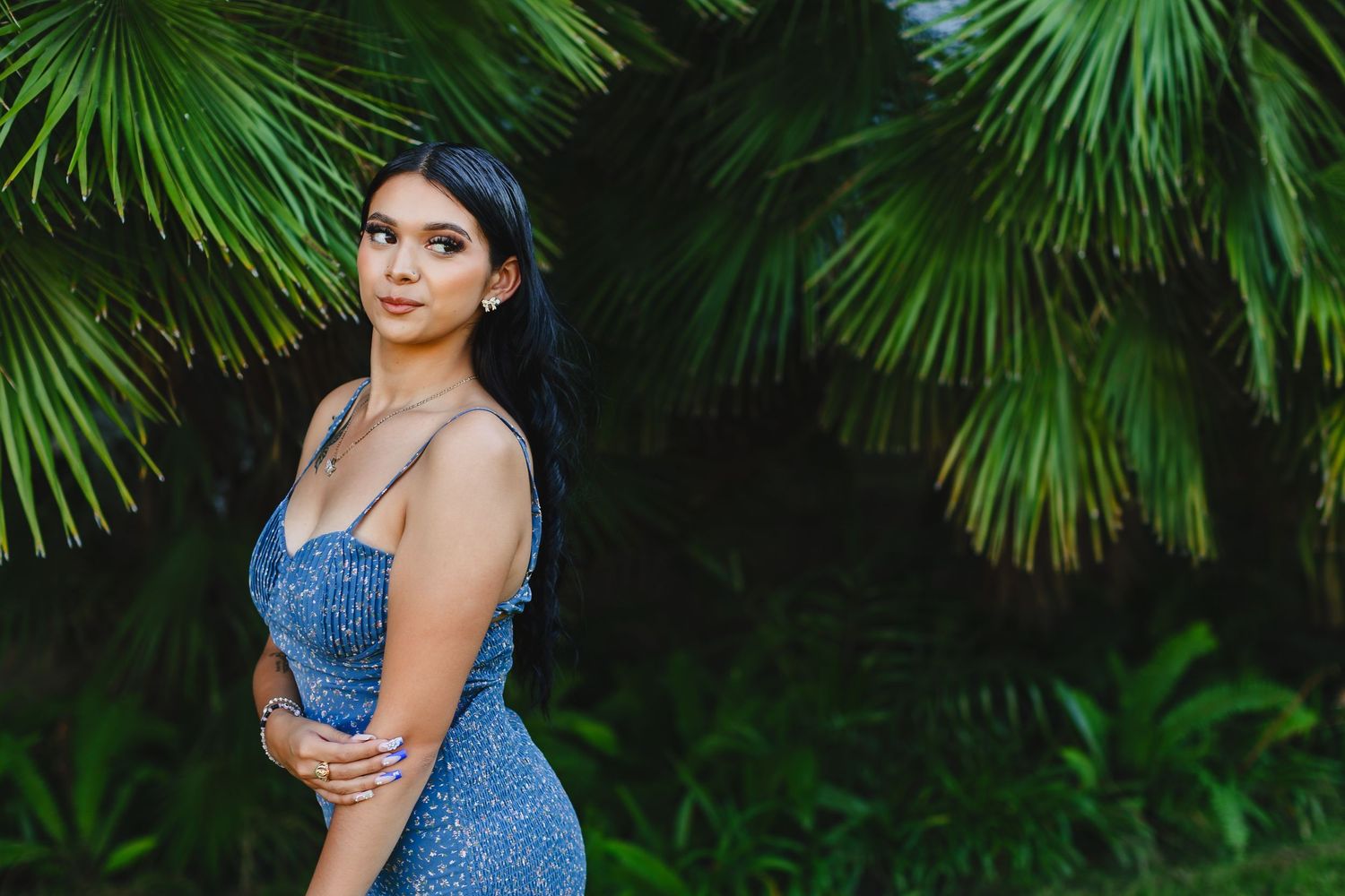 Woman in sparkly blue dress poses against tropical palm frond background in outdoor portrait setting.