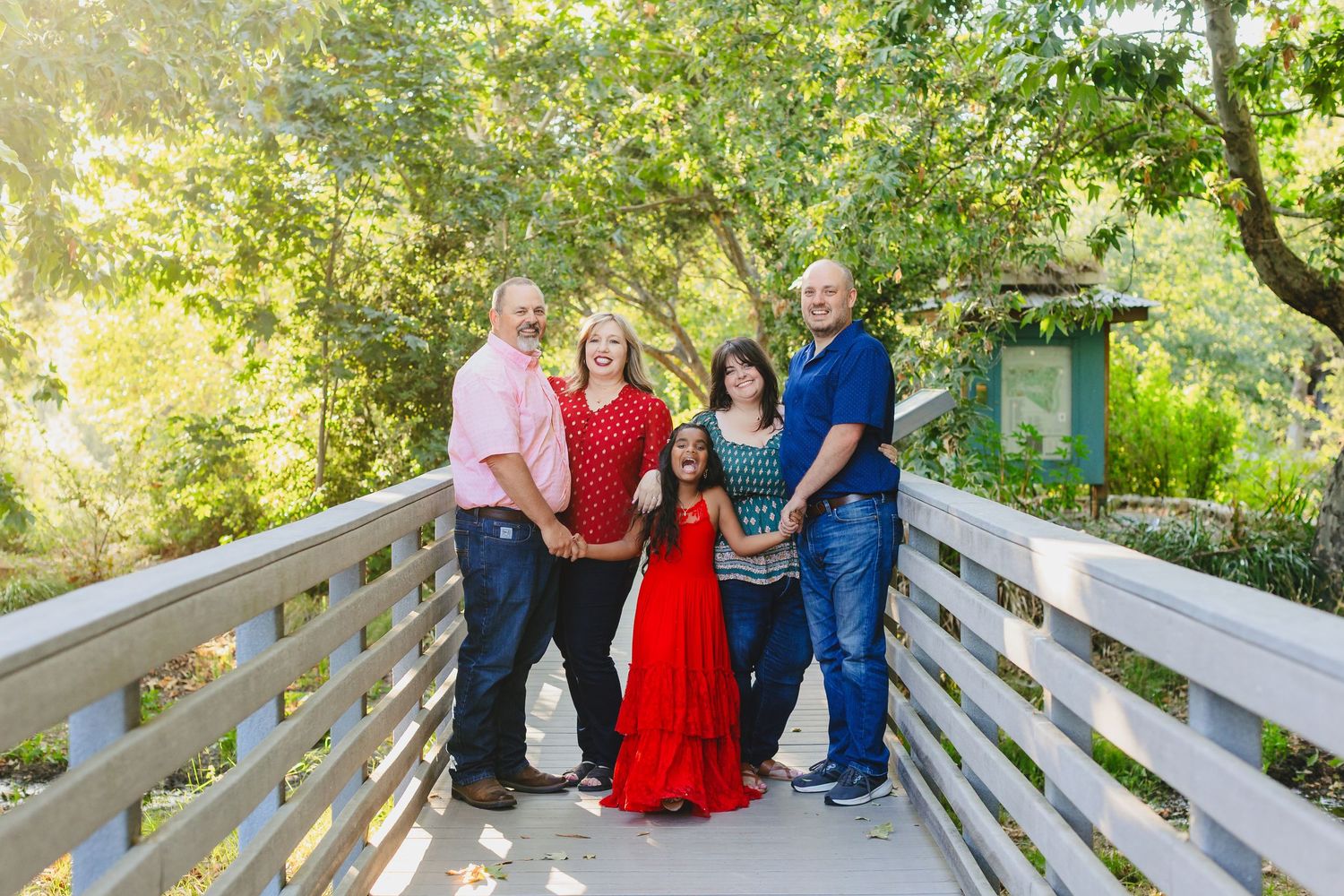 A group poses together on a wooden bridge walkway surrounded by lush green trees on a sunny day.