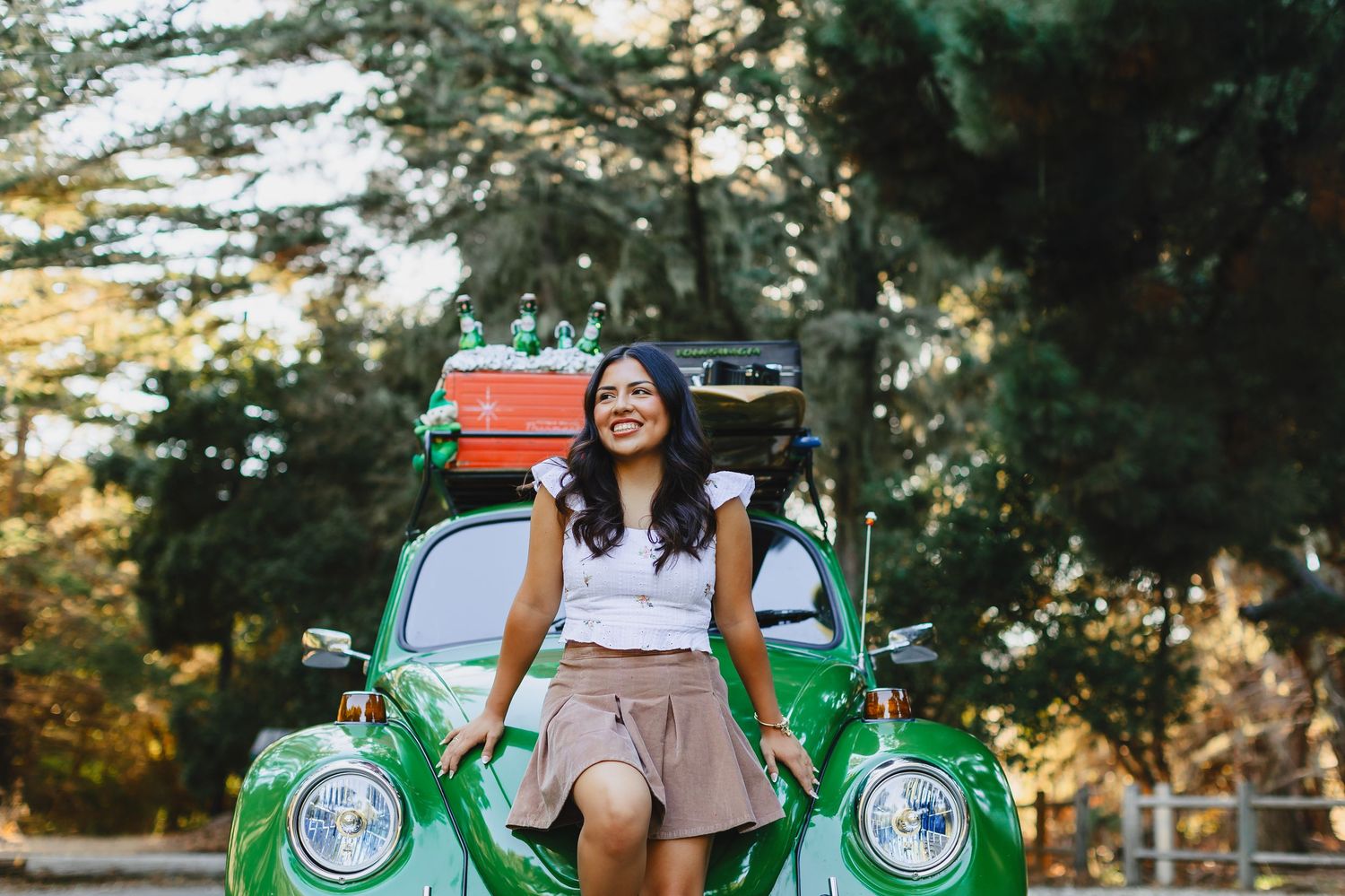 Smiling young woman leaning on a vintage green VW Beetle with luggage on top outdoors.
