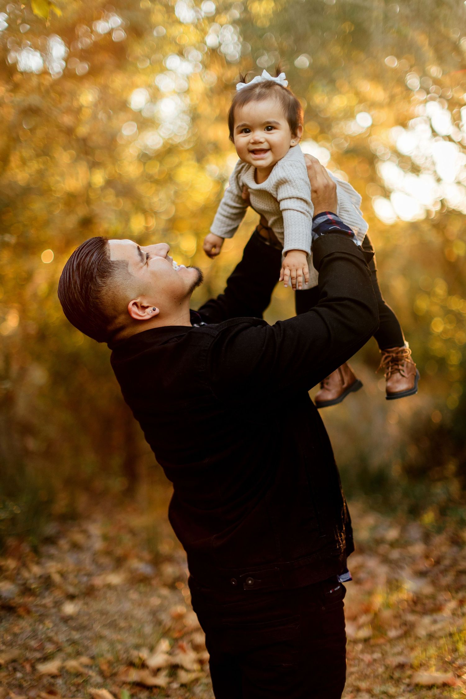 Adult in black sweater lifts small child in air among autumn leaves during golden sunset play time outdoors.
