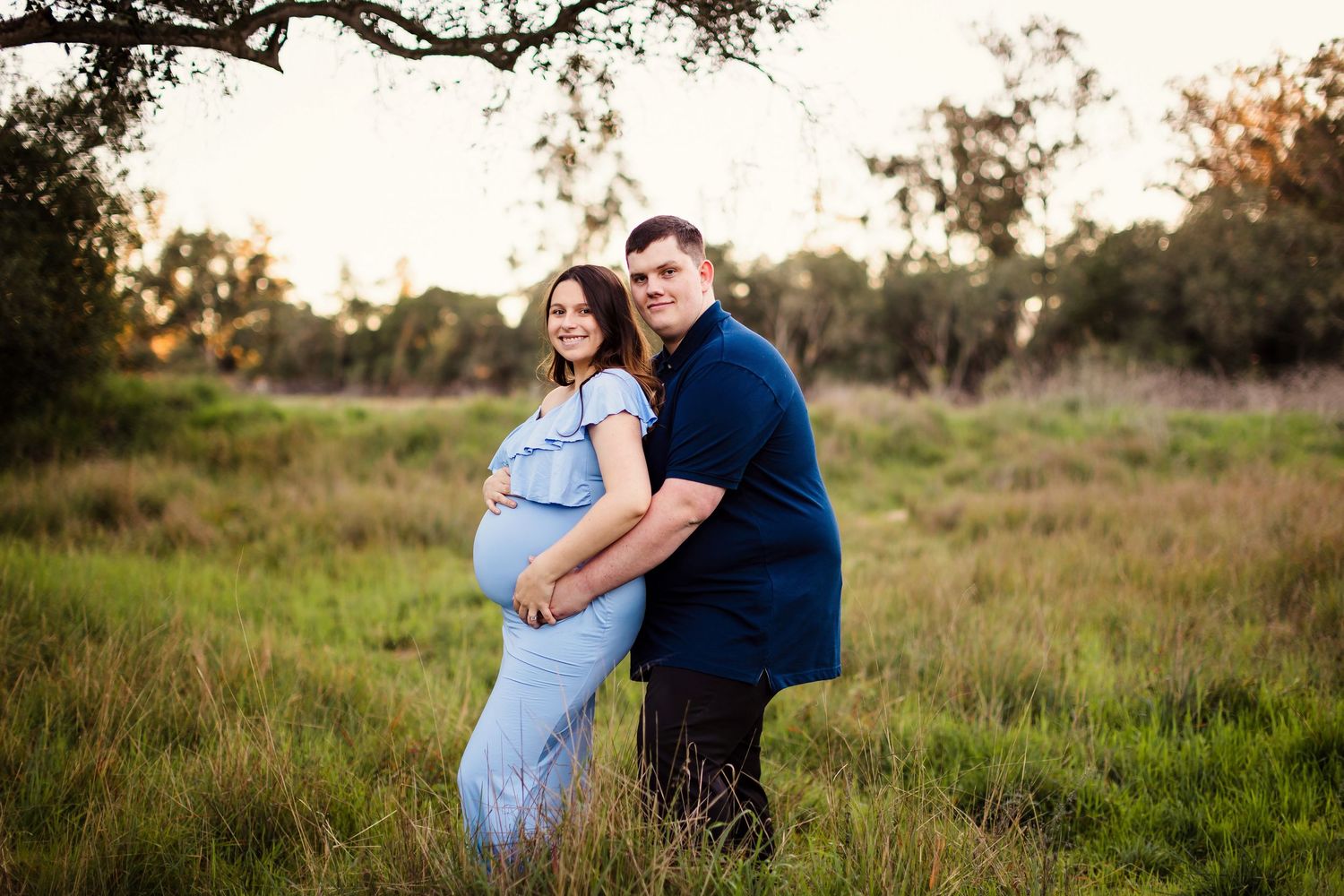 A romantic pregnancy photoshoot shows tender moments between a couple in a lush grassy field at sunset.