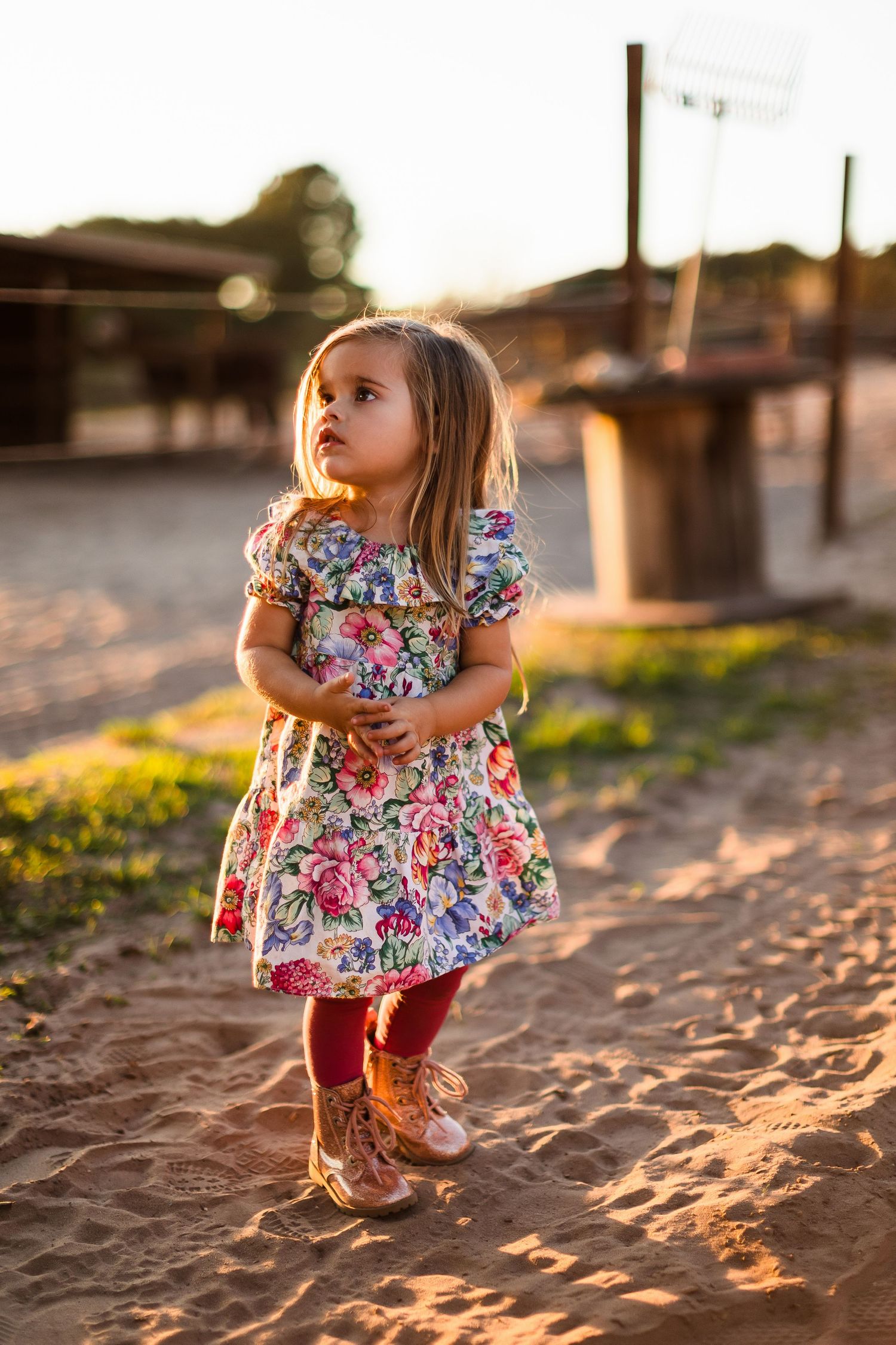 A young child in a floral dress and boots stands on sandy ground during golden hour sunset lighting.