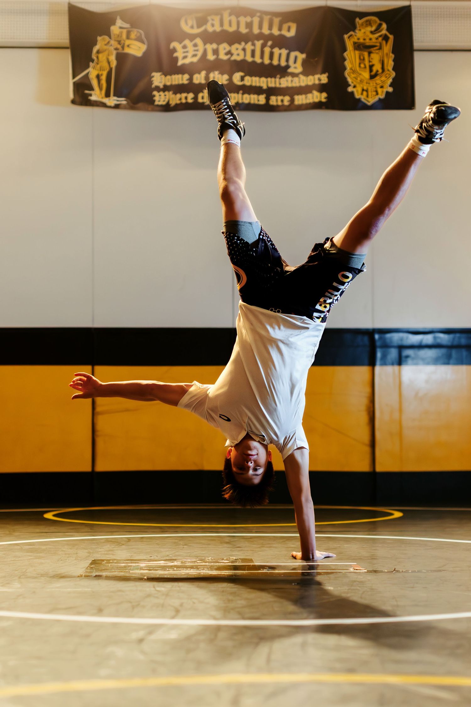 Wrestler performs a one-handed handstand on a wrestling mat at Cabrillo Wrestling gym, Home of the Conquistadors.
