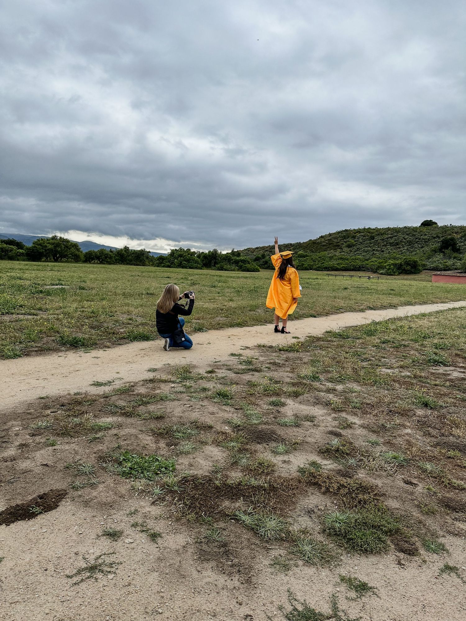 Photographer captures graduate in yellow cap and gown posing on dirt path in grassy field.