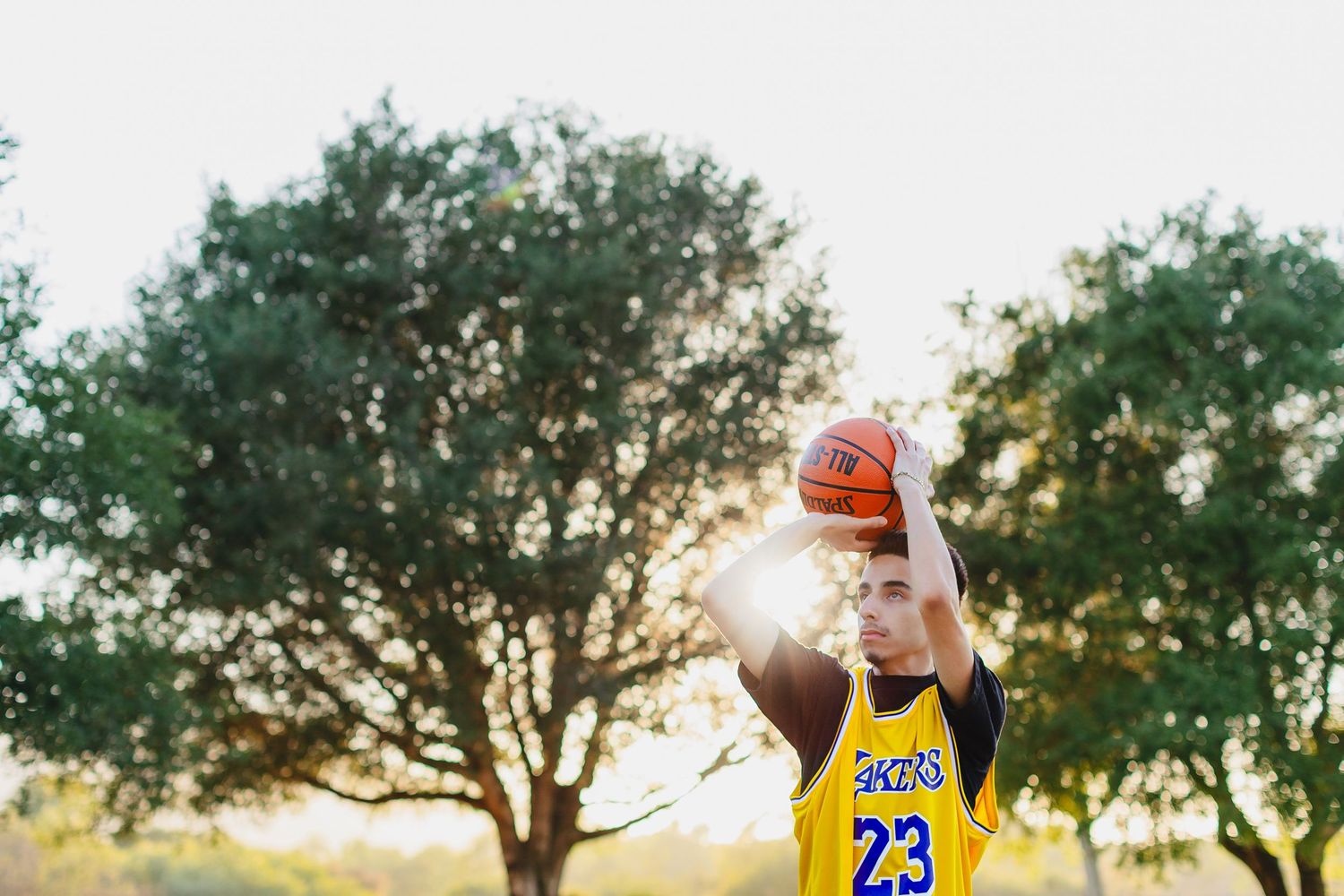 Young basketball player in yellow jersey number 23 holding ball overhead outdoors with sunlight through trees.