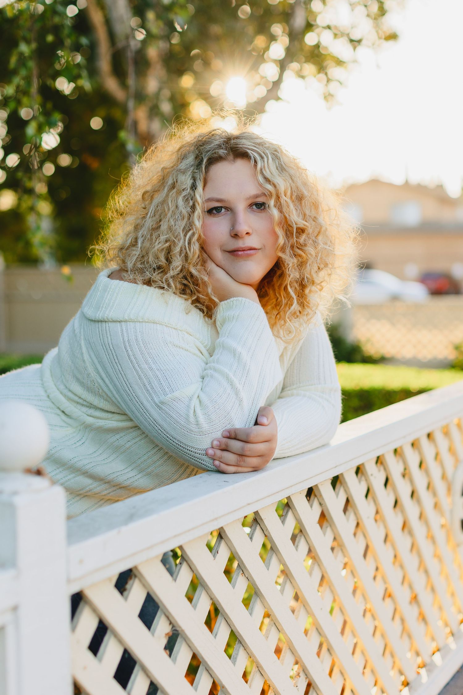 Person with curly blonde hair in white sweater leaning on white fence during sunset.