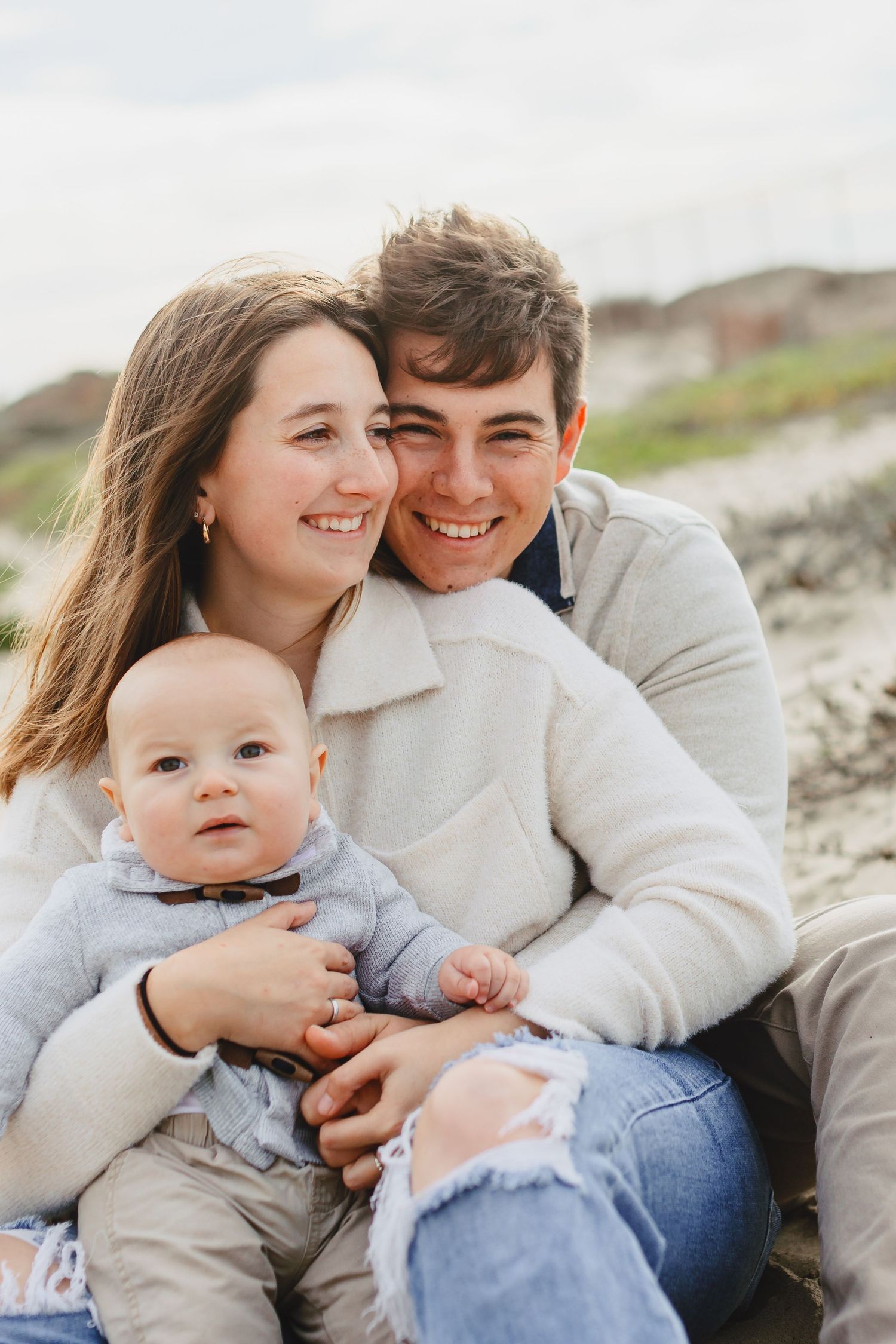 A young family smiles together while holding their baby during a beach photoshoot.