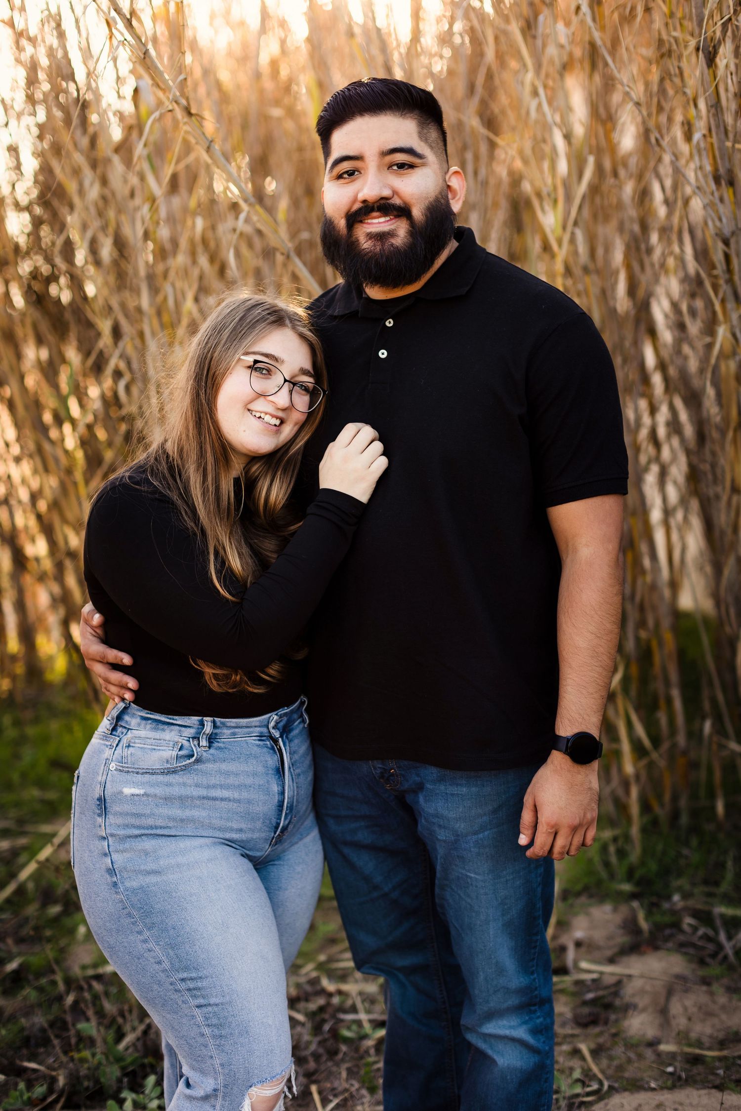 A couple poses intimately in a sunset field of tall reeds, with warm golden lighting creating a romantic atmosphere.