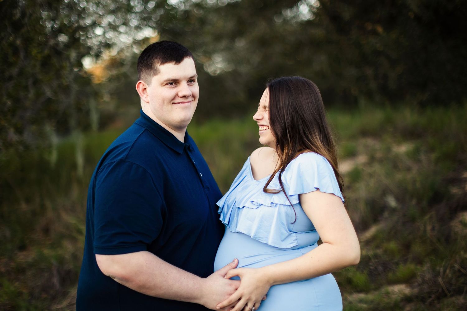 A couple in blue outfits share a tender moment in a natural outdoor setting during maternity photoshoot.