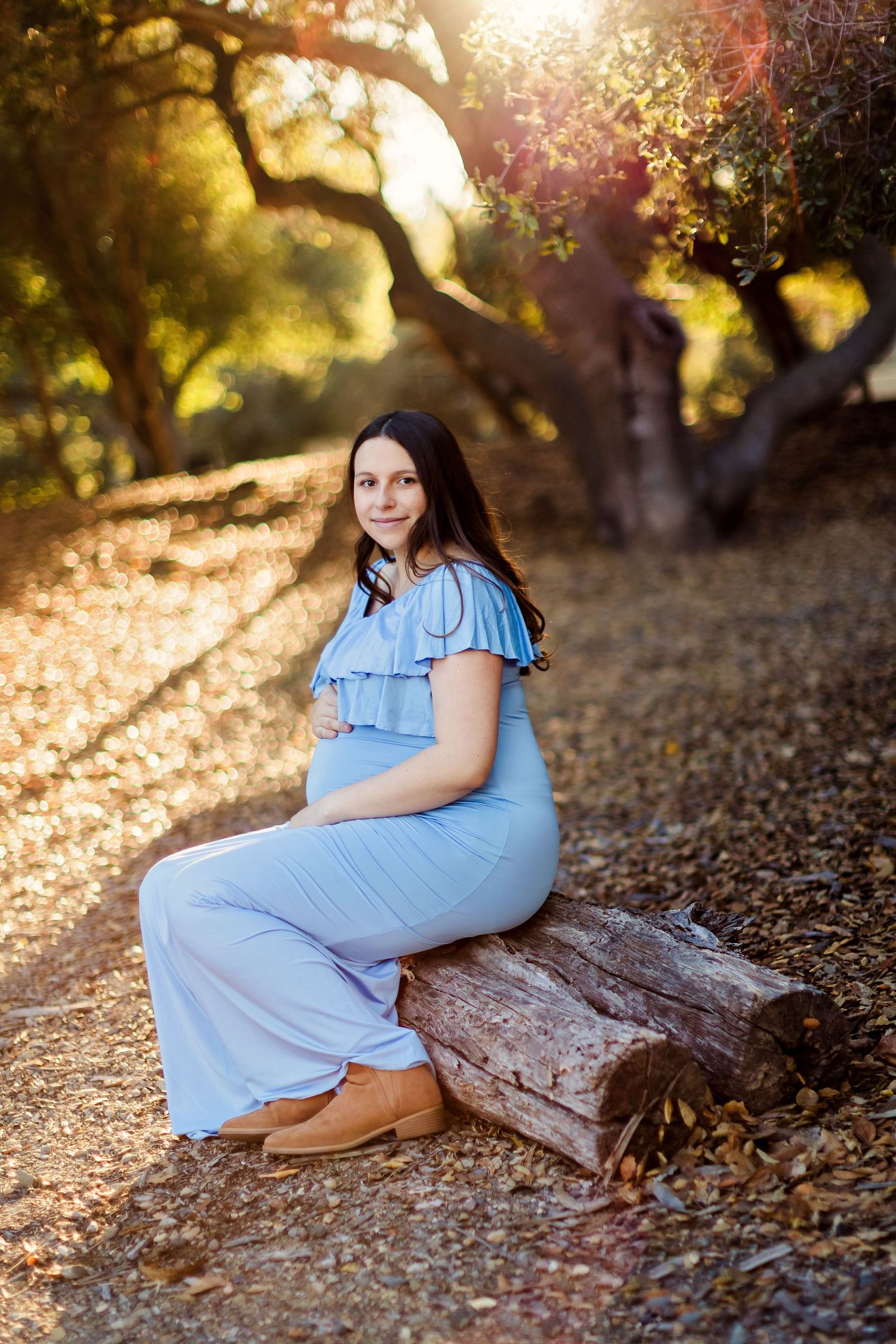 Four serene maternity photos taken in a golden sunlit forest with fallen autumn leaves and rustic logs.