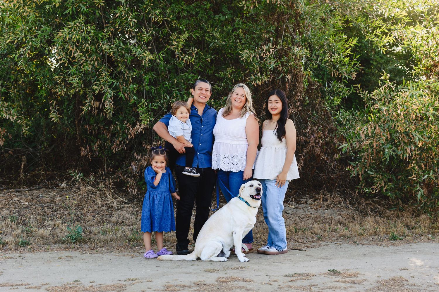Family of four with a white Labrador dog posing outdoors near lush green trees on a sandy path.