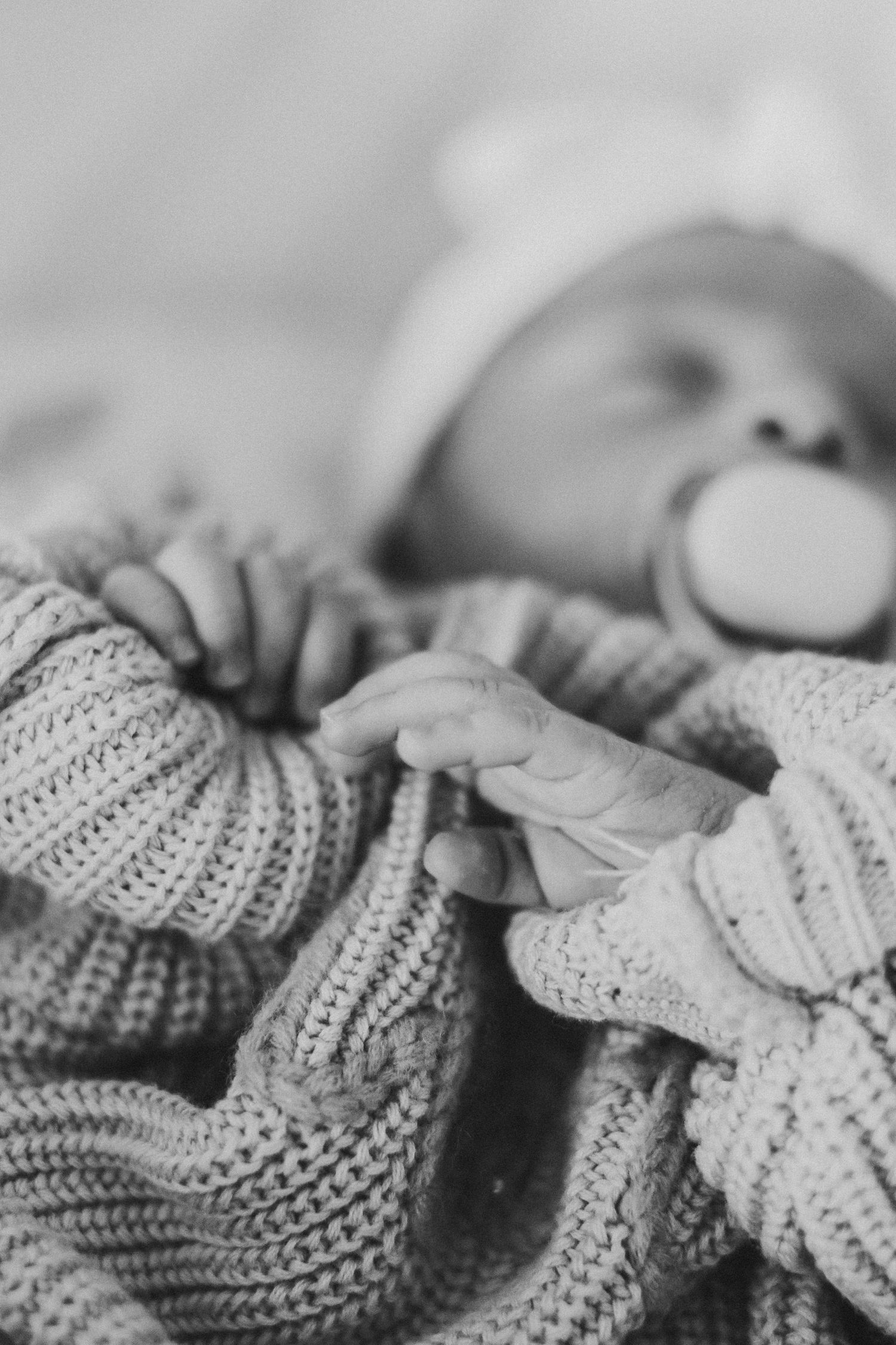 Black and white photo of a newborn baby with a pacifier, wrapped in a knit blanket, tiny hands folded together.