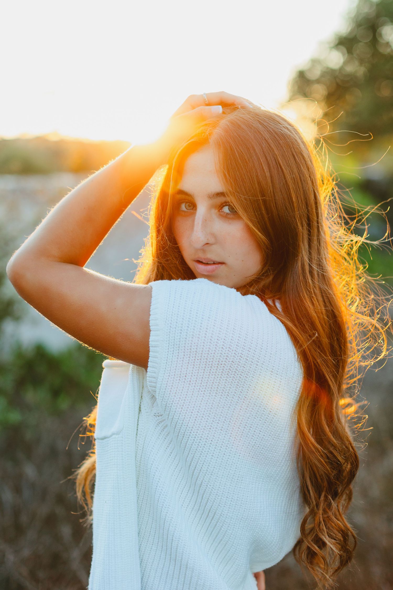 Young woman with long red hair in white sweater poses outdoors at golden sunset hour.