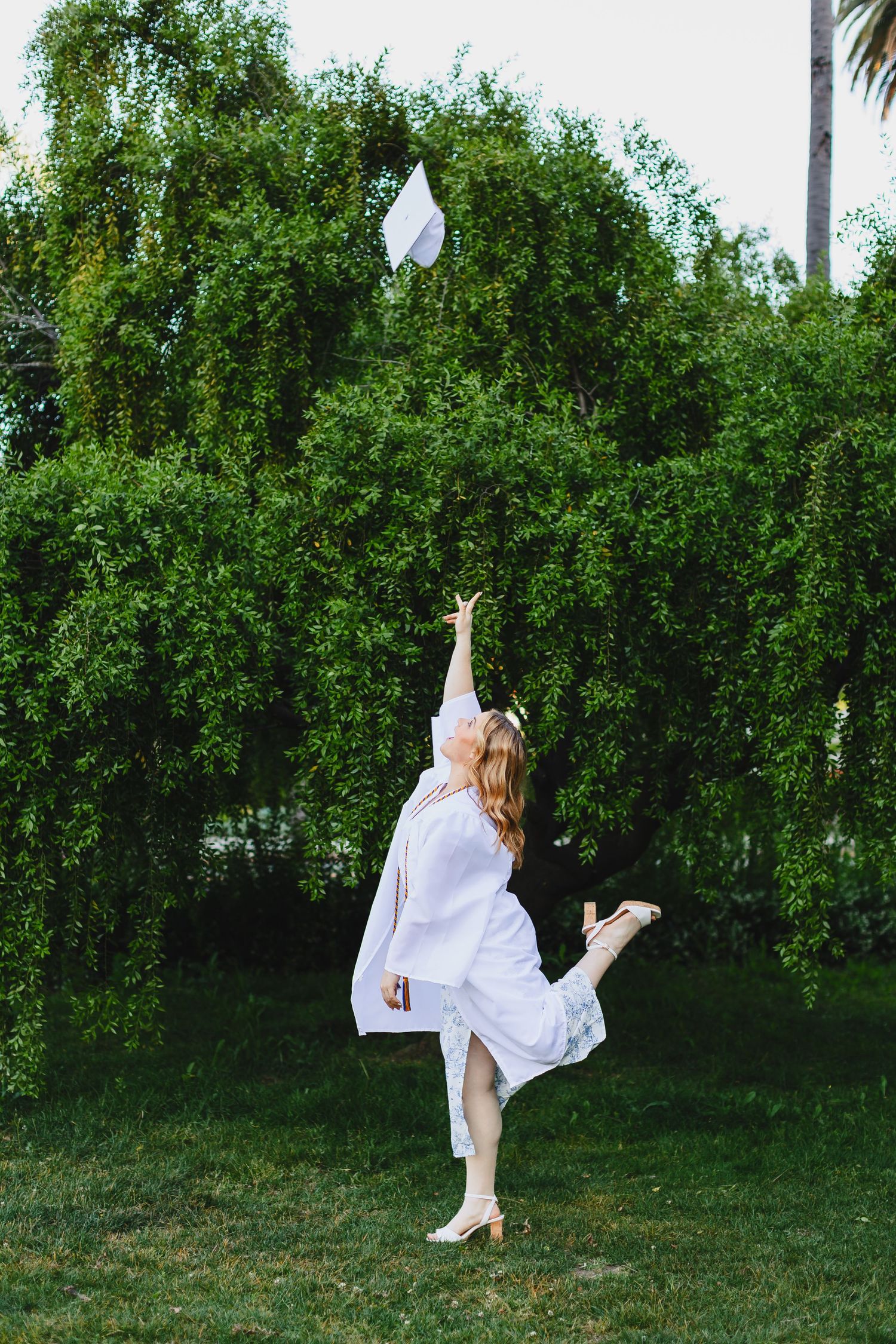 Woman in white dress throwing graduation cap in lush garden setting.