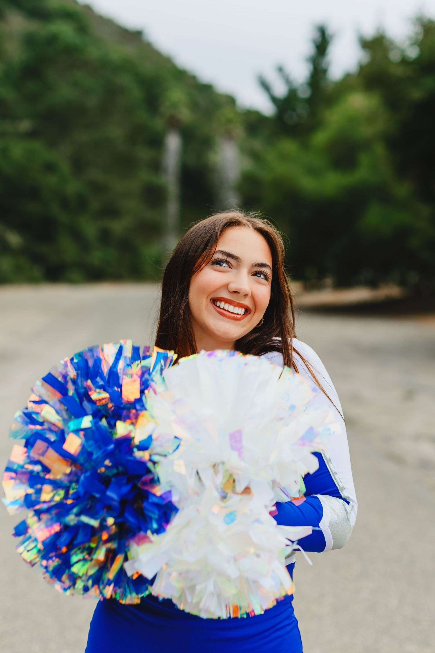 Cheerleader in royal blue outfit holding white and blue pom poms while smiling outdoors.