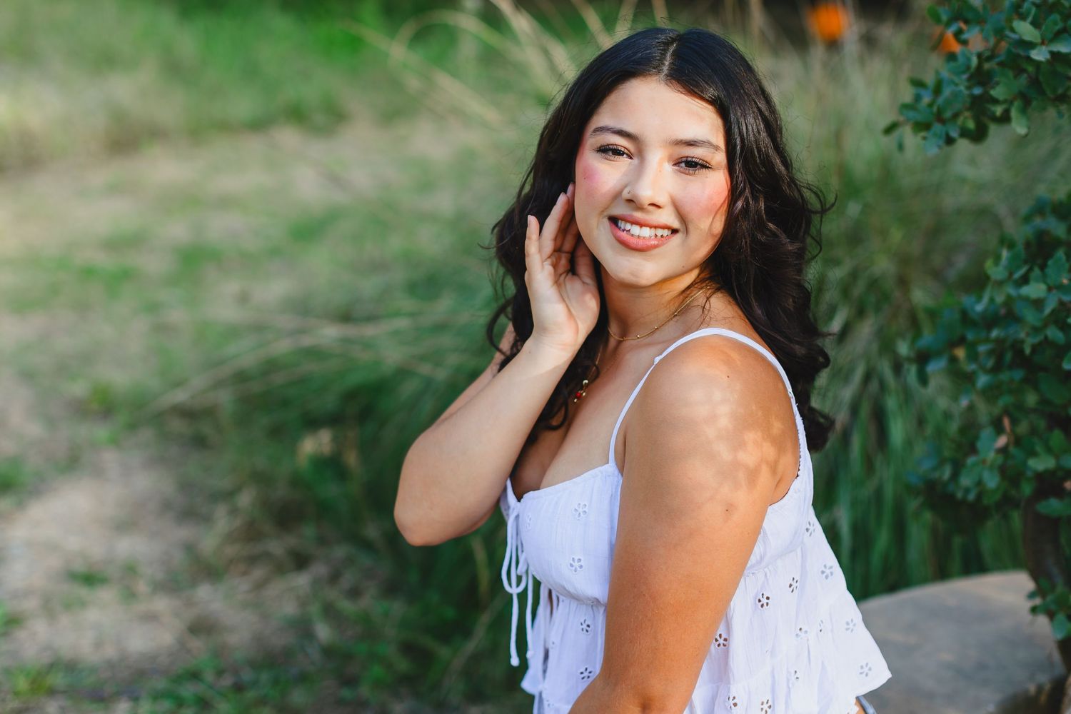 A person with dark hair and a white tank top smiles warmly near green foliage on a sunny day.
