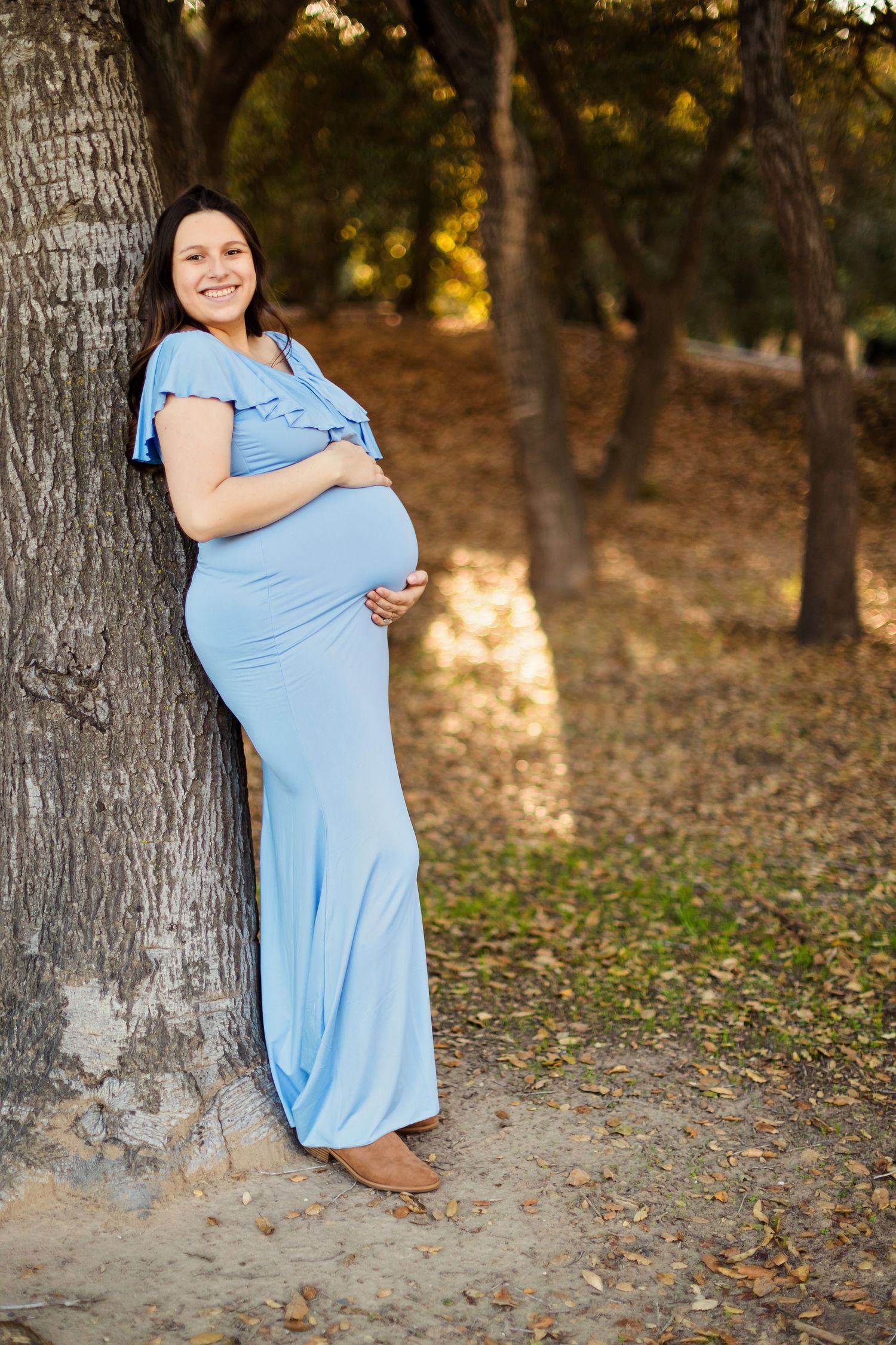 A pregnant woman in a light blue dress poses in a natural outdoor setting with autumn leaves and trees.