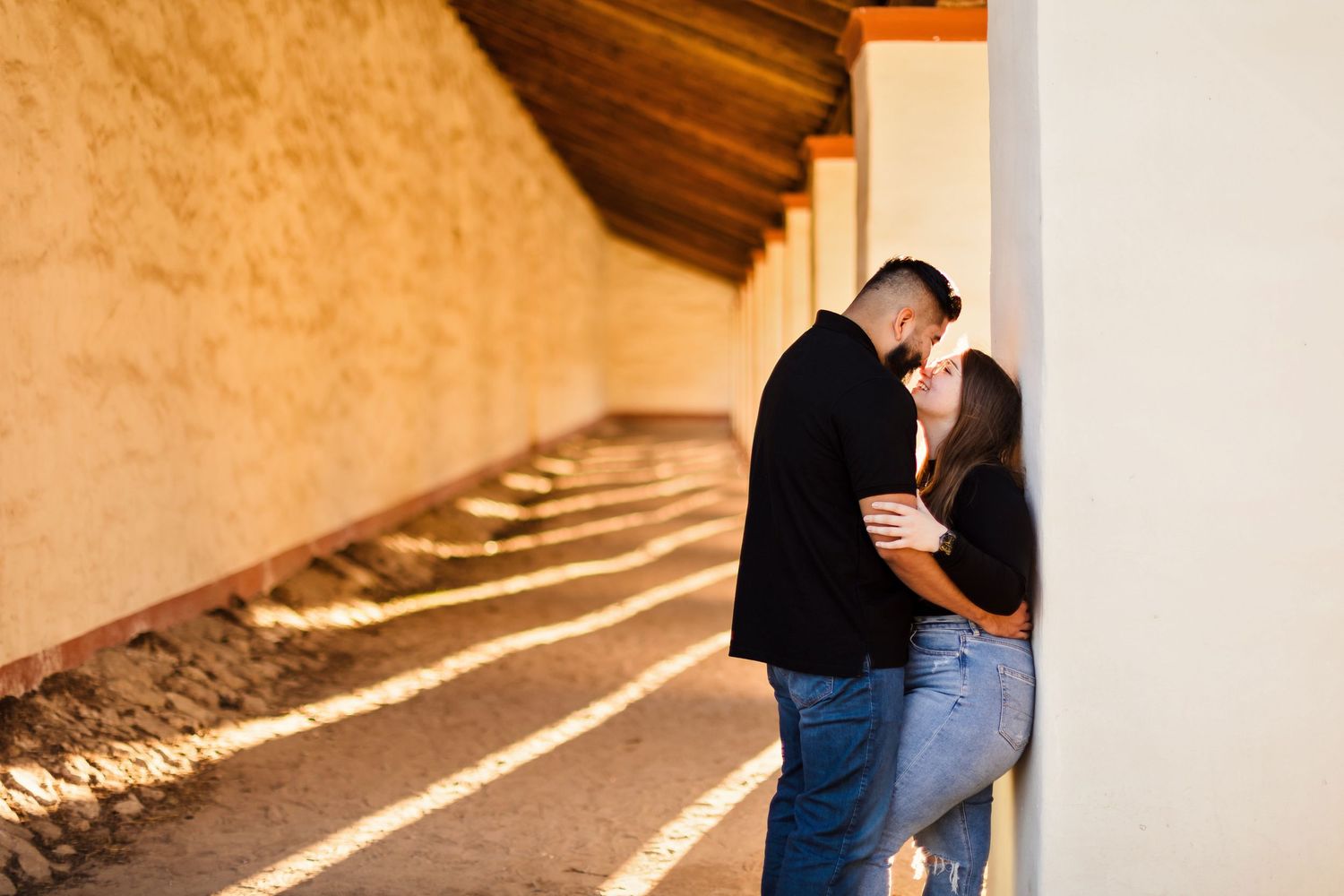 A couple shares an intimate moment in a sunlit colonnade with white walls and terracotta tones.