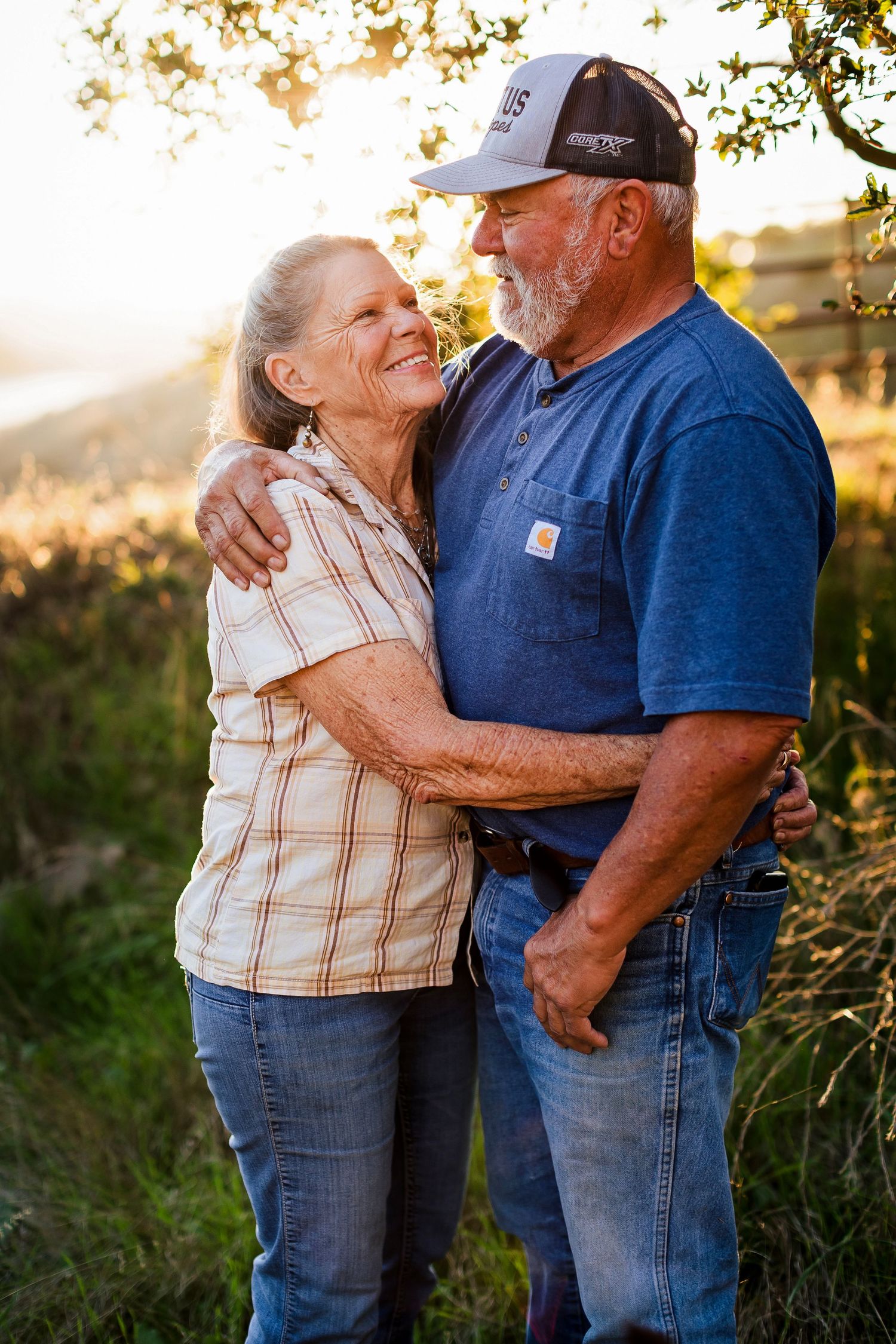 An elderly couple shares a tender embrace in a golden sunset field, wearing casual jeans and plaid clothing.