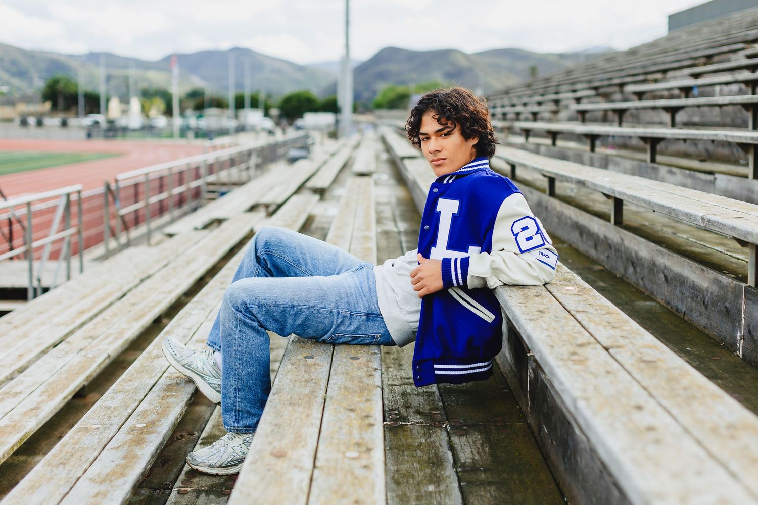 Person in blue varsity jacket and jeans lying across concrete stadium bleachers in outdoor setting.
