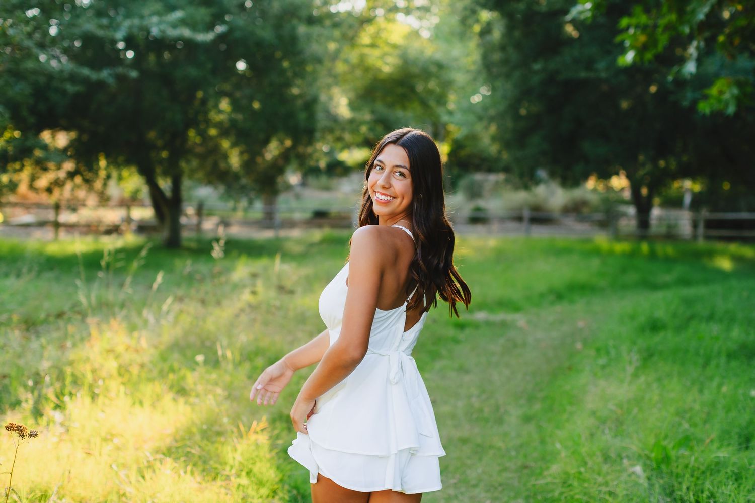 Smiling brunette woman twirling in a white dress in a sunny green field.