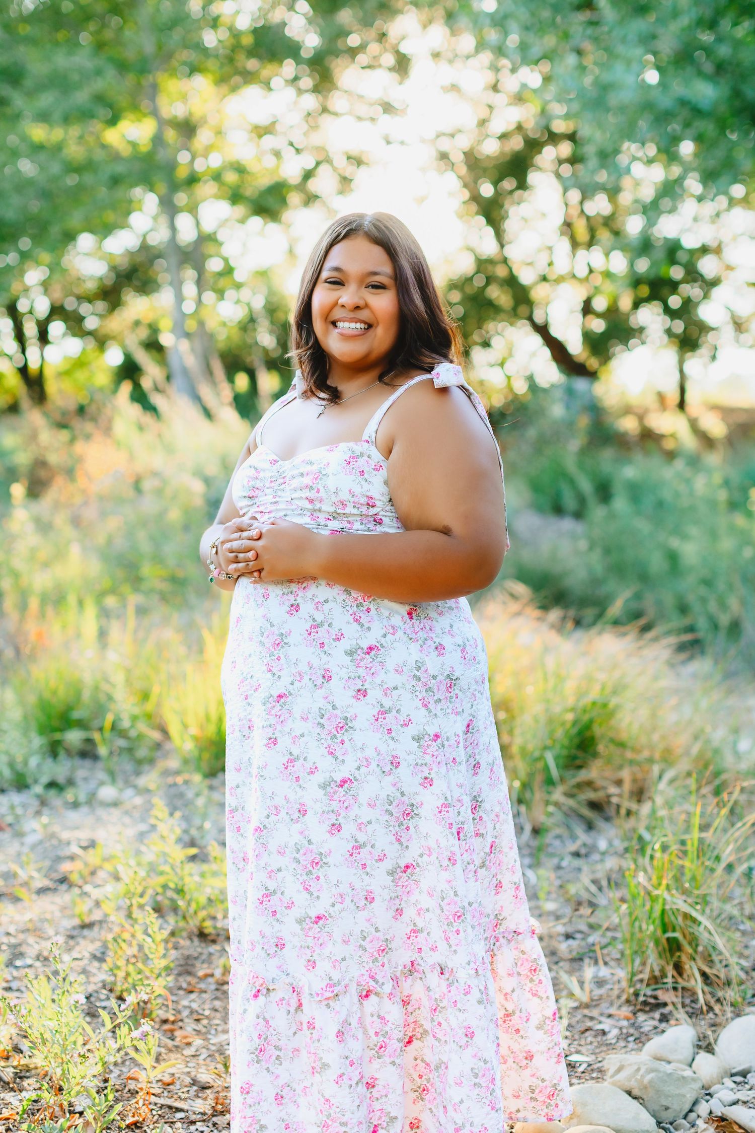 Smiling young woman in white floral maxi dress posing outdoors in a sunlit garden path.