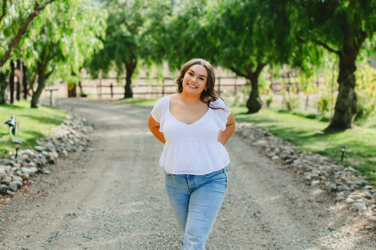 Someone walks happily down a tree-lined dirt path on a sunny day wearing jeans and a white top.