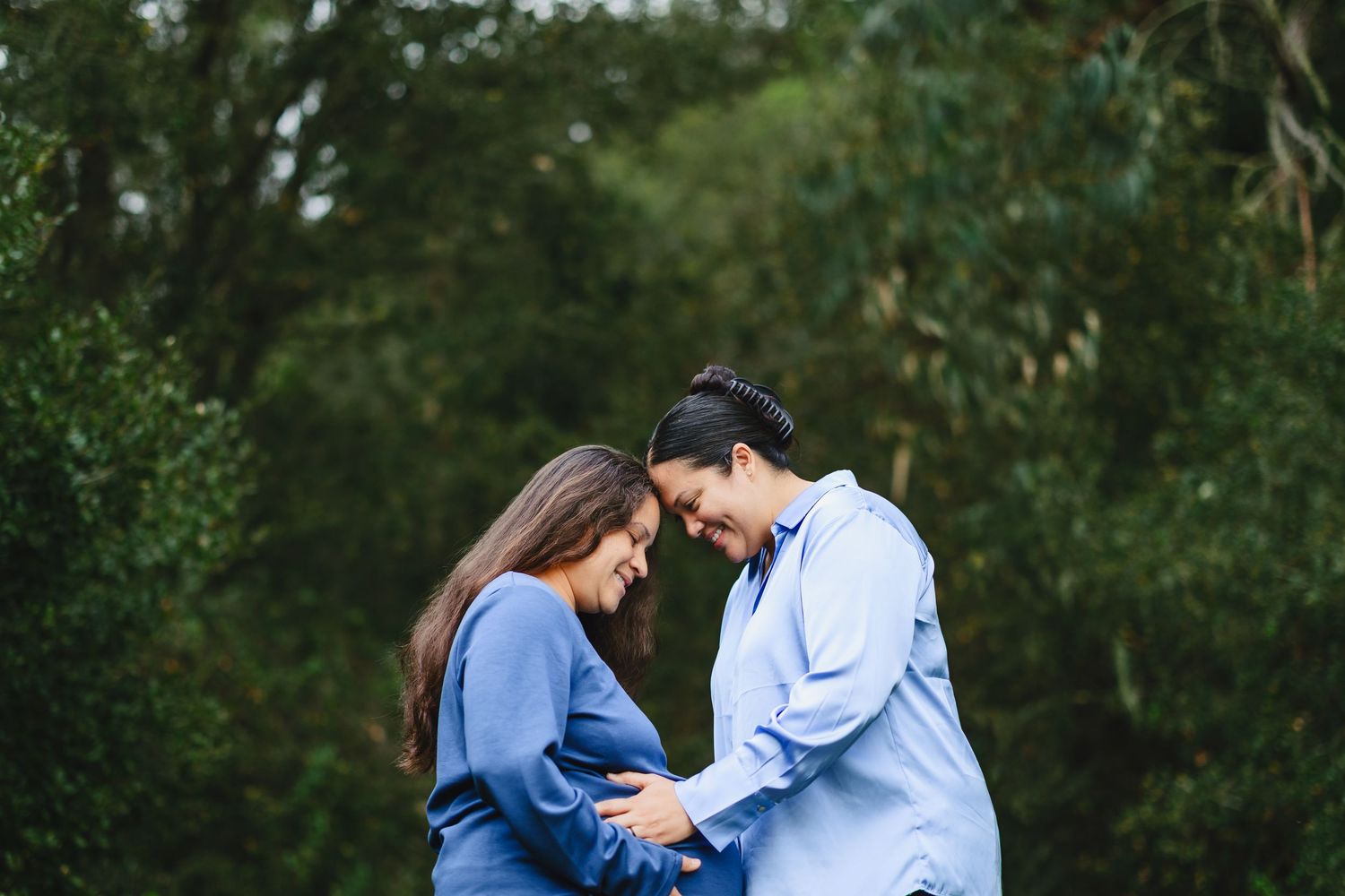 Couple in blue outfits sharing a tender moment outdoors, heads bowed together over a baby bump, surrounded by lush green trees.