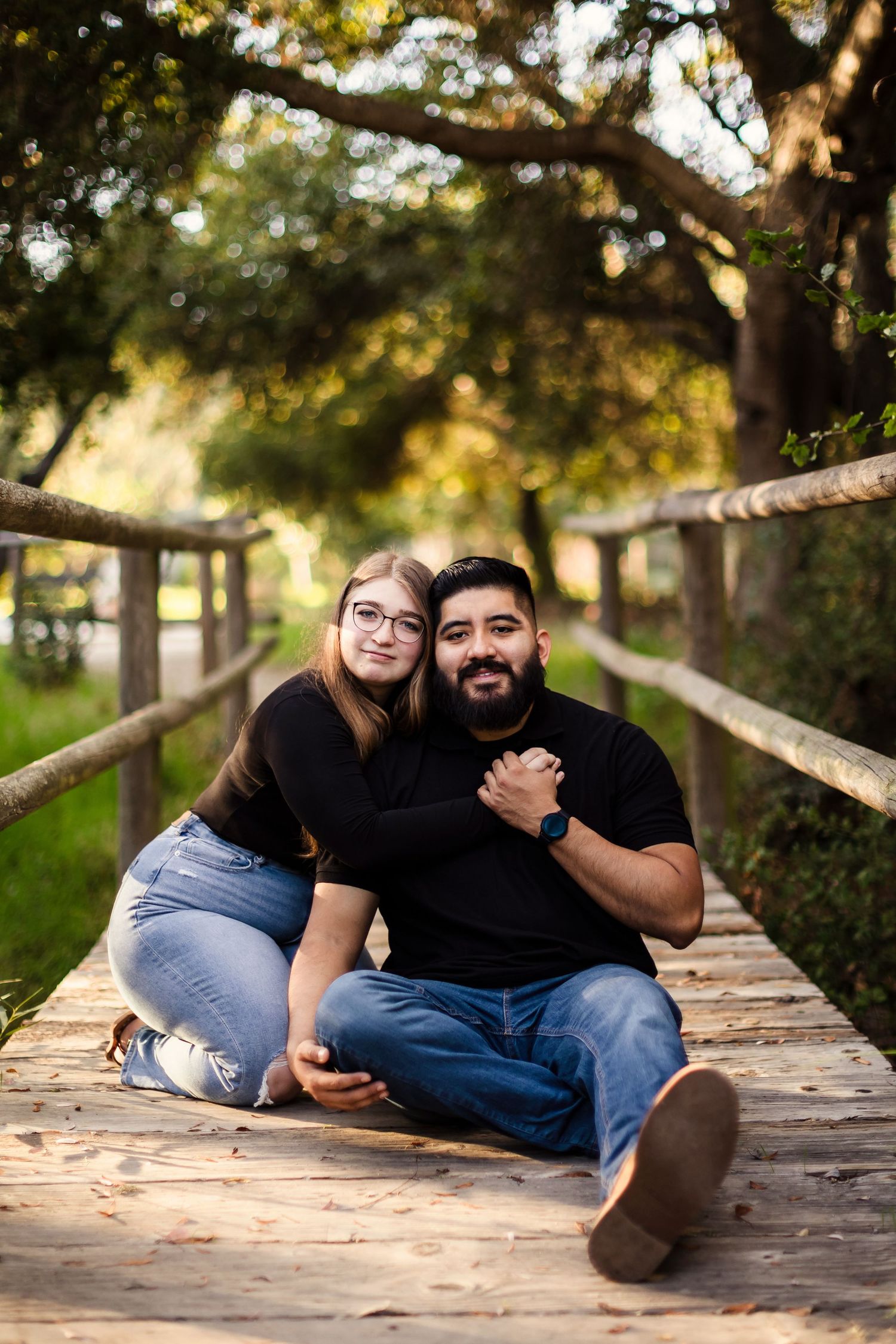 A loving couple sits and reclines on a wooden boardwalk surrounded by trees in golden afternoon light.