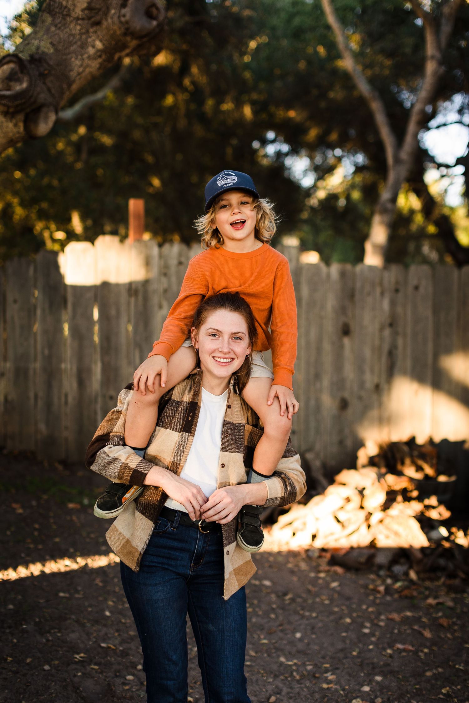 A child wearing an orange sweater sits on another person's shoulders in front of a wooden fence at sunset.