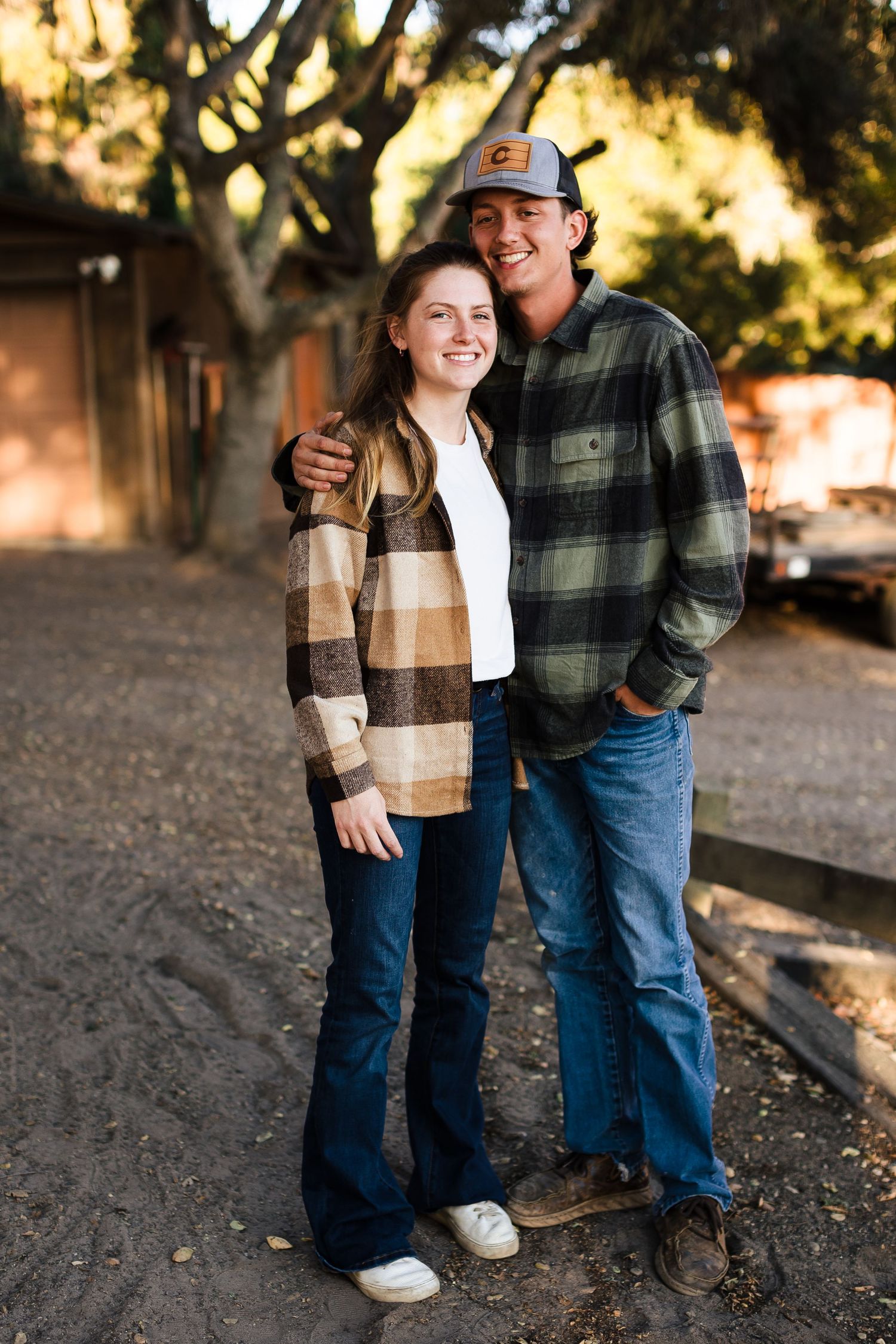 A couple wearing plaid flannel shirts and jeans pose together outdoors at sunset on a dirt path.
