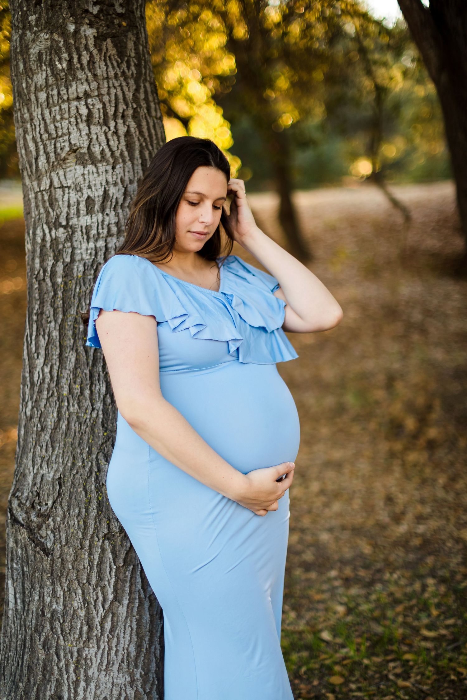 A pregnant woman in a light blue ruffled dress poses thoughtfully among autumn trees and fallen leaves.