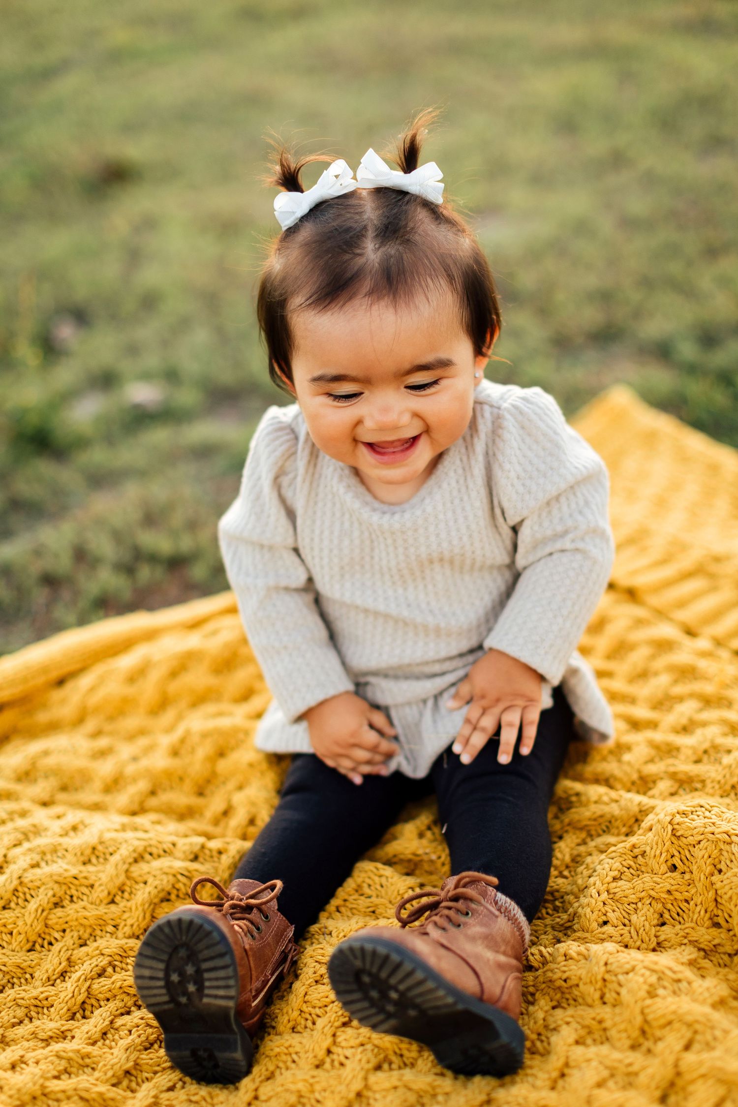 A child sits on a yellow blanket outdoors wearing a gray sweater and leather shoes while smiling and looking down.