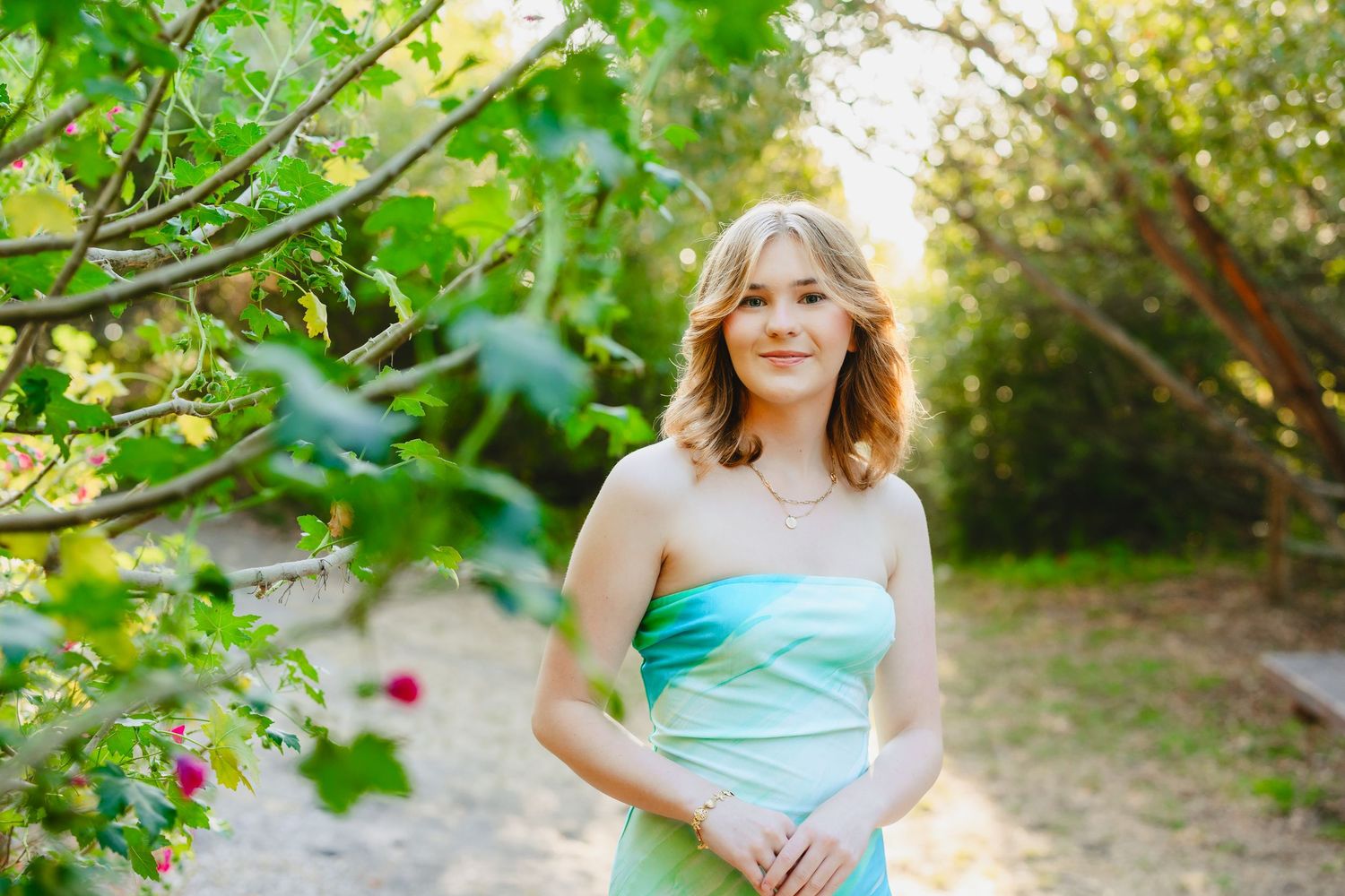 Young woman in a teal strapless dress posing outdoors among lush green trees on a sunny day.