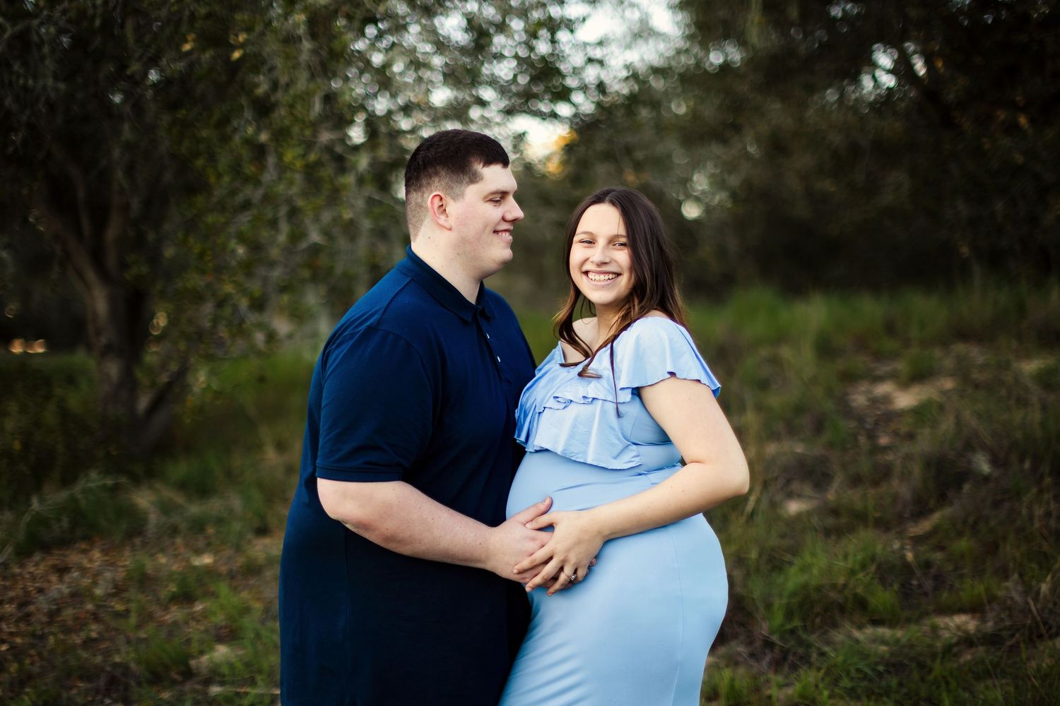 A romantic couple embraces and laughs together during an outdoor photoshoot in a wooded setting.