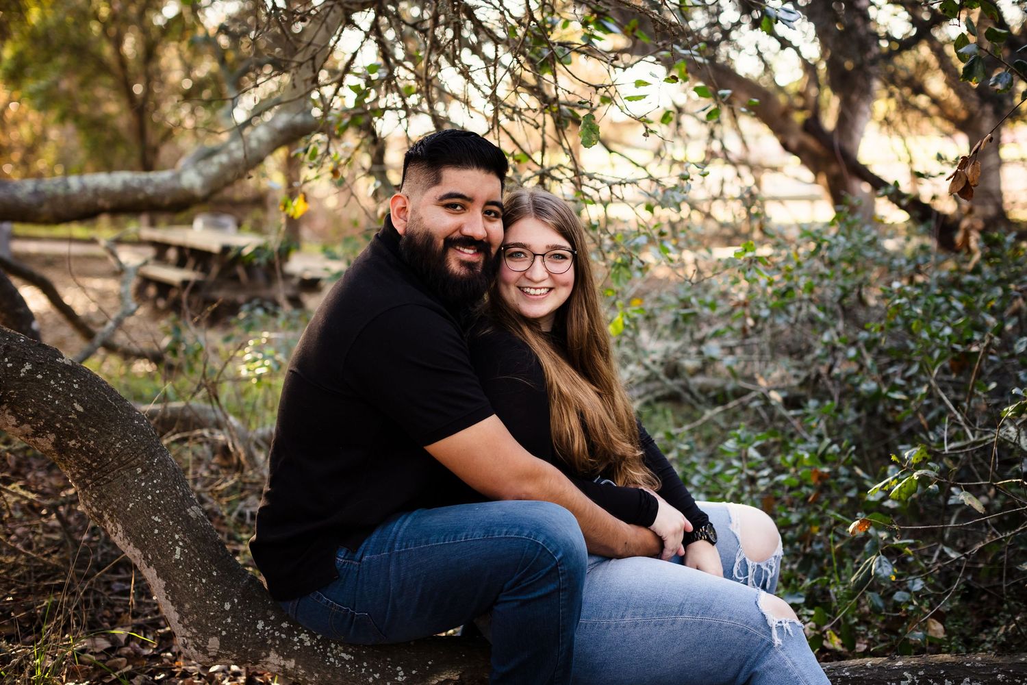 A couple sitting together outdoors in a natural setting surrounded by trees and foliage on an autumn day.