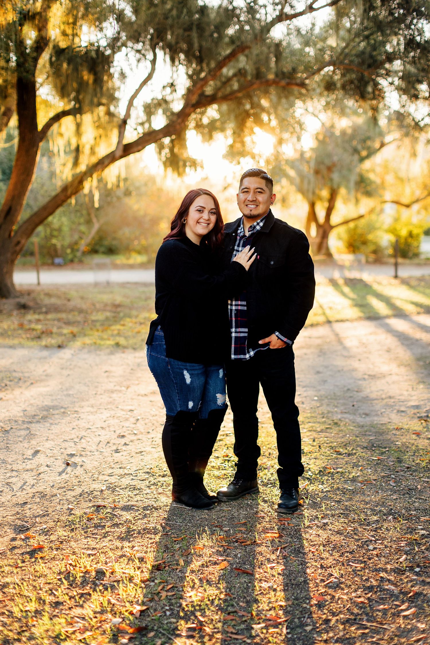 A pair stand together outdoors during golden hour sunlight filtering through trees on an autumn path.