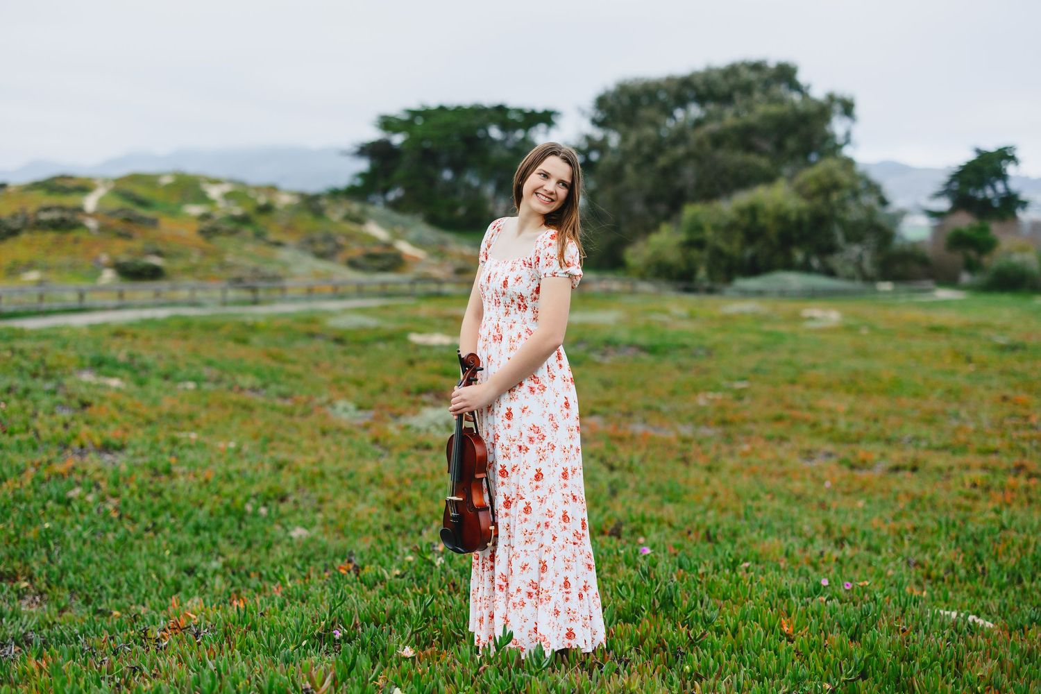 A smiling woman in a floral maxi dress holds a violin while standing in a lush green meadow with trees in the background.