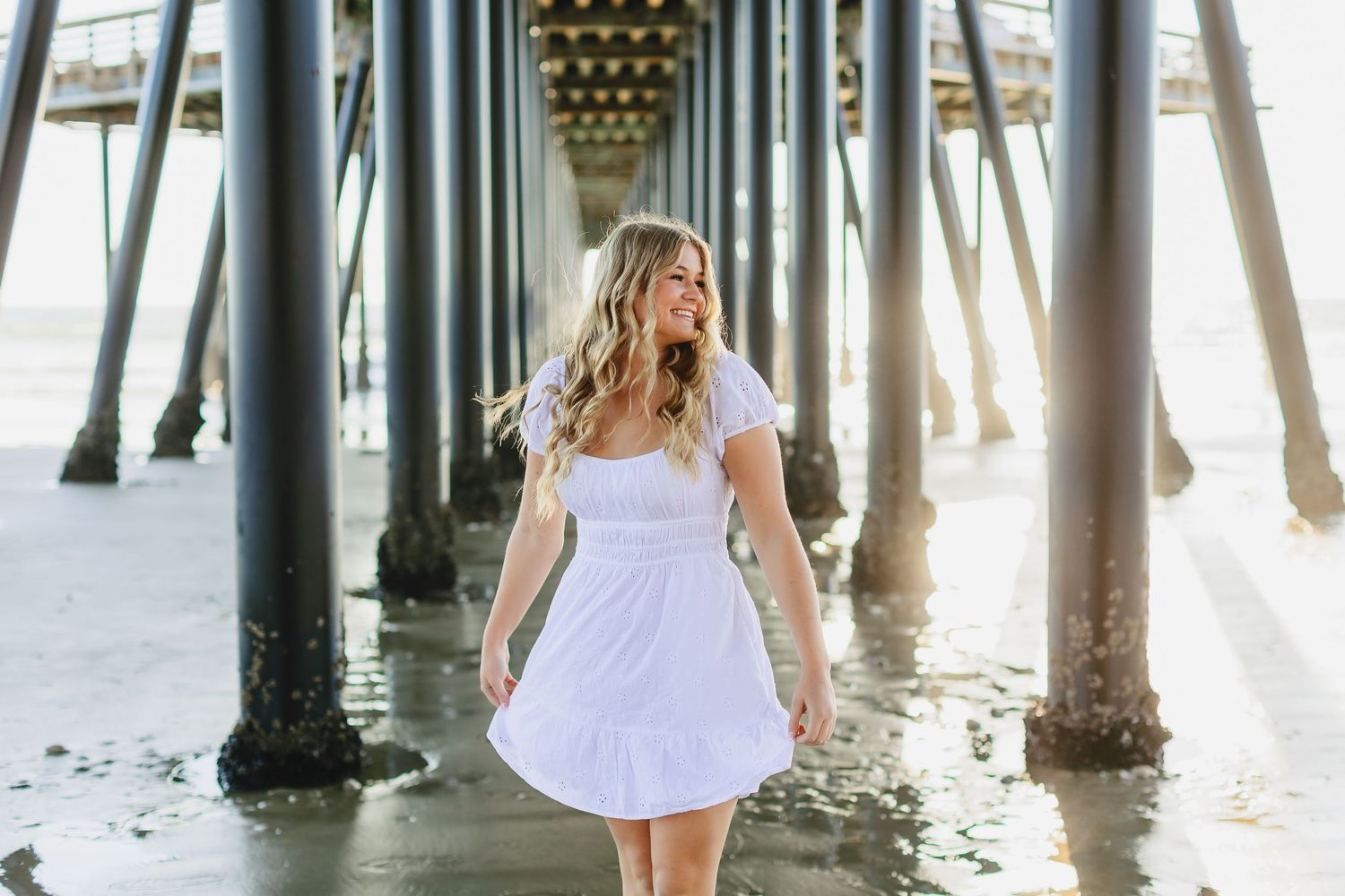 Someone in a white summer dress stands beneath a wooden pier while sunlight streams through the pillars at the beach.