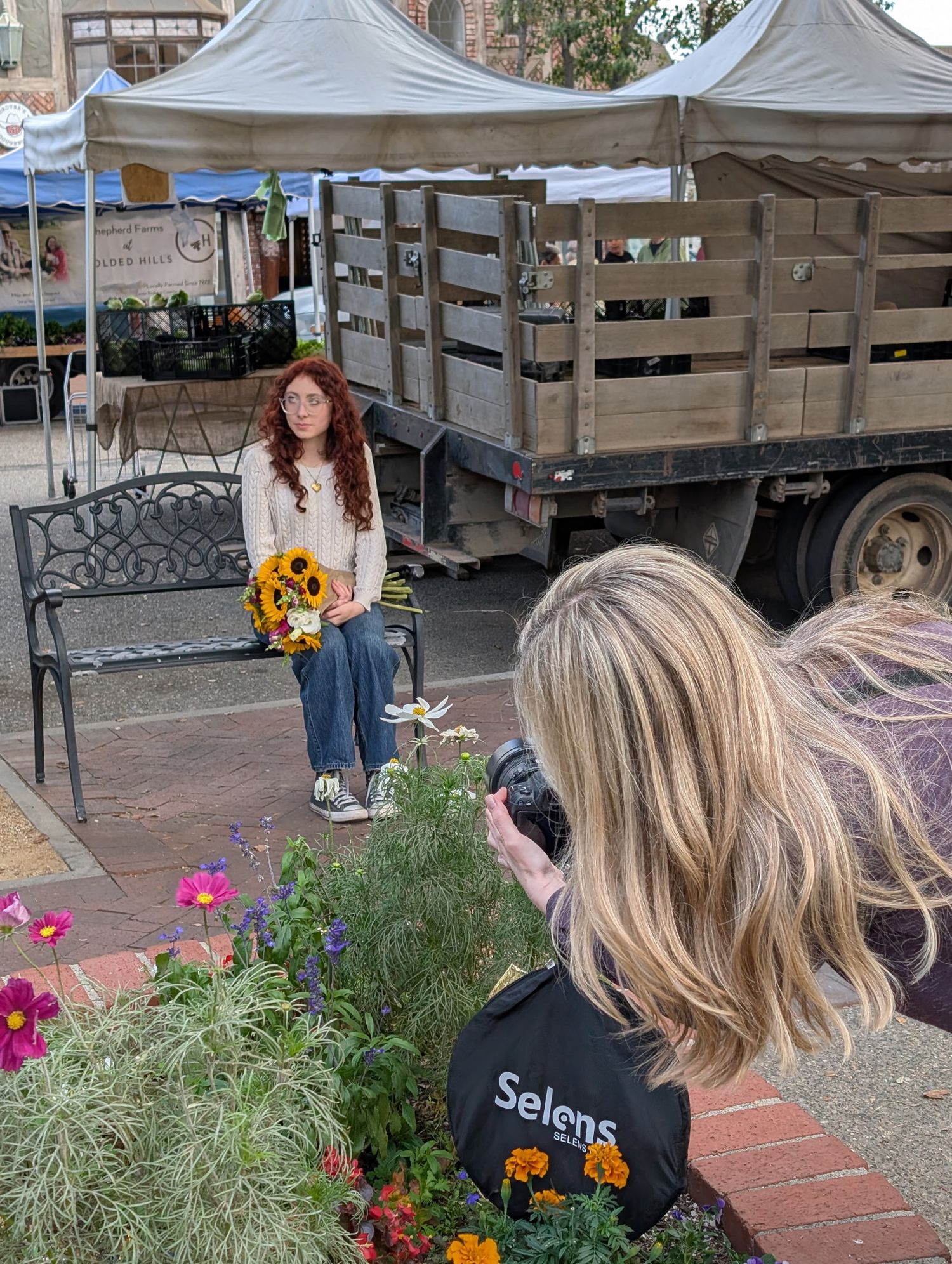 Photographer photographing a redhead woman holding sunflowers at an outdoor farmers market.