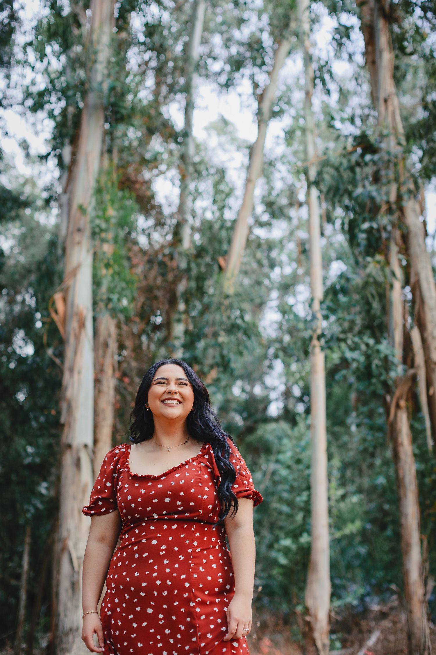 Young woman in red floral dress smiling among tall eucalyptus trees in outdoor portrait.