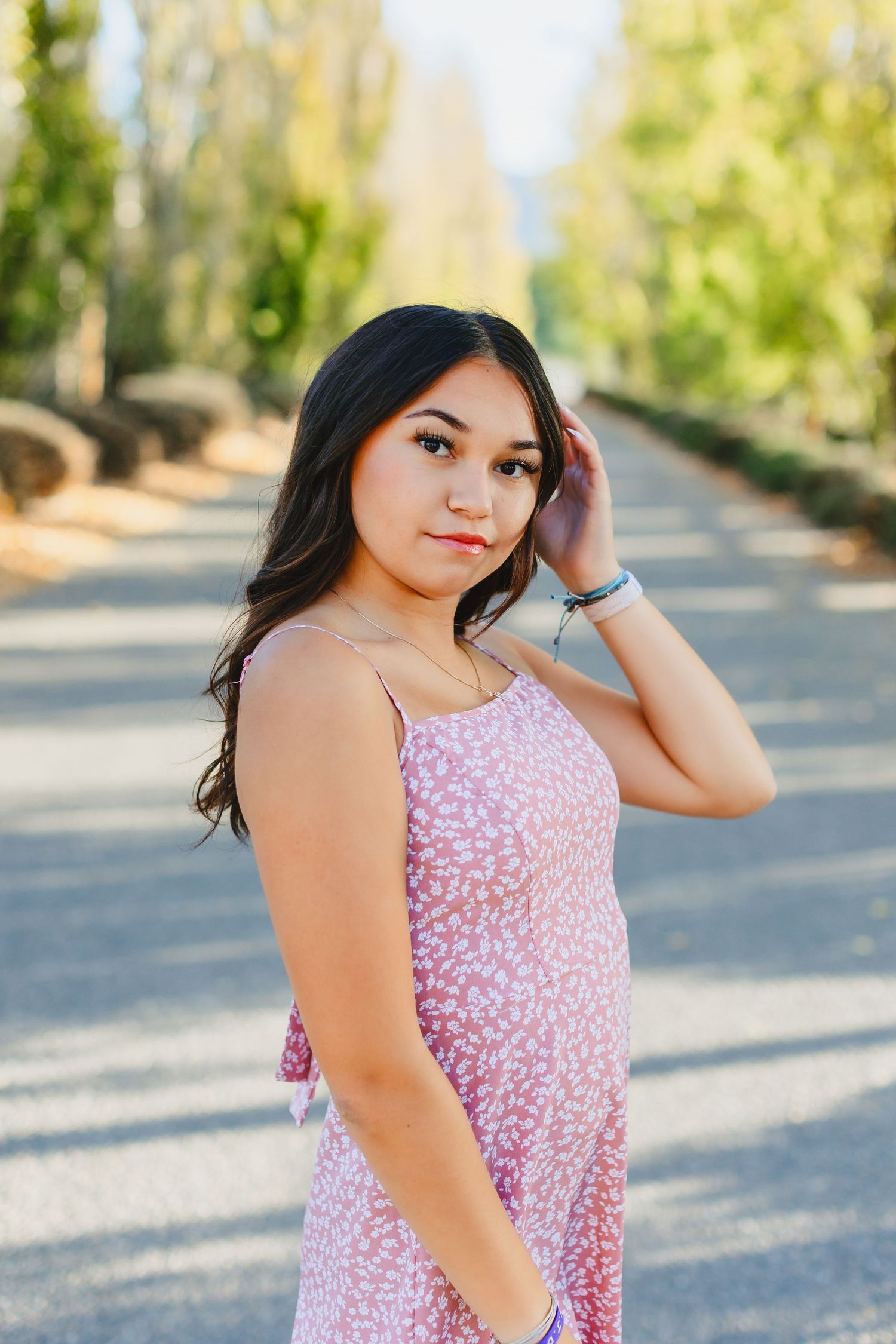 Teen girl in pink floral sundress stands on a tree-lined path during golden hour sunlight.