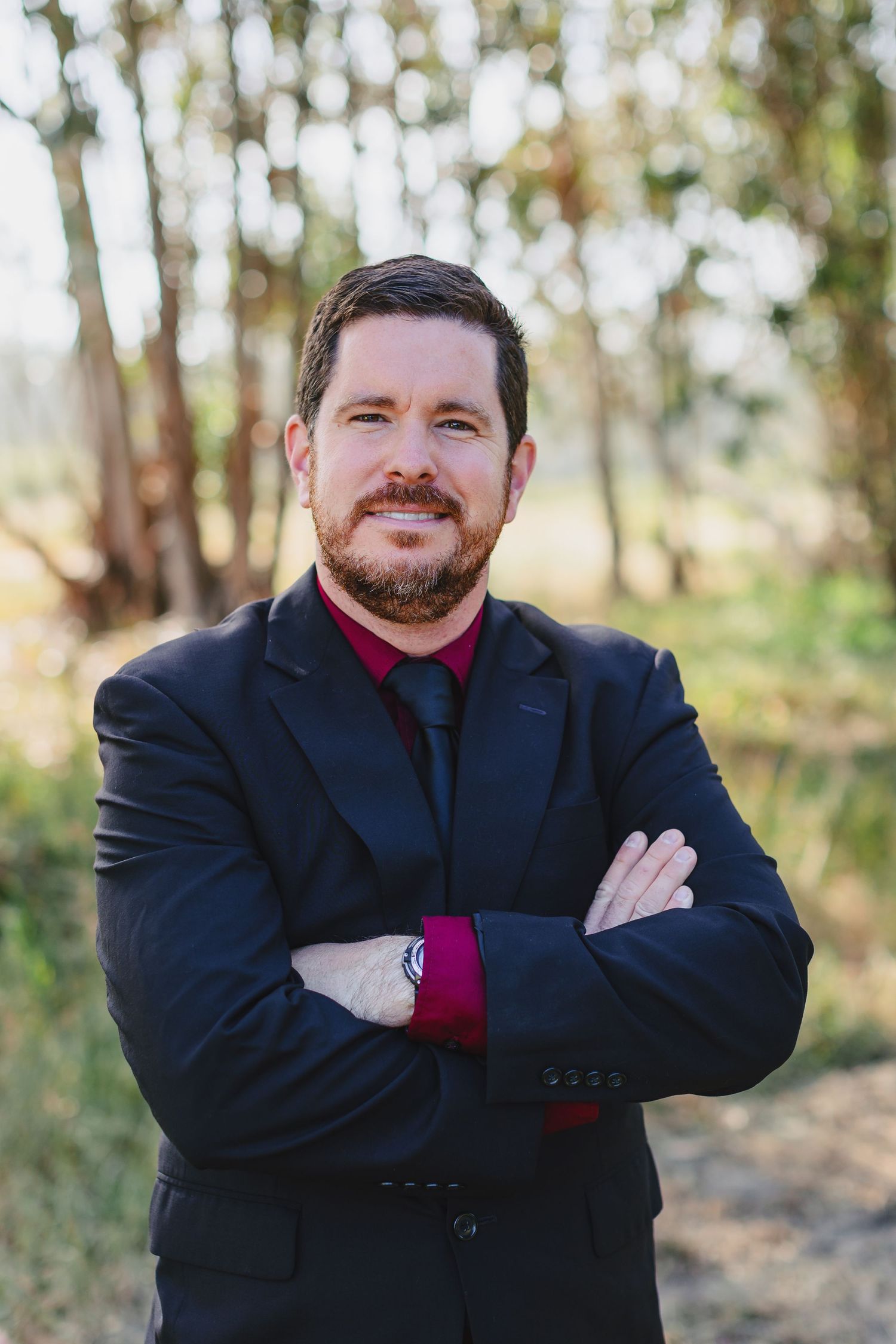 Professional in black suit and red shirt standing with crossed arms against natural backdrop.