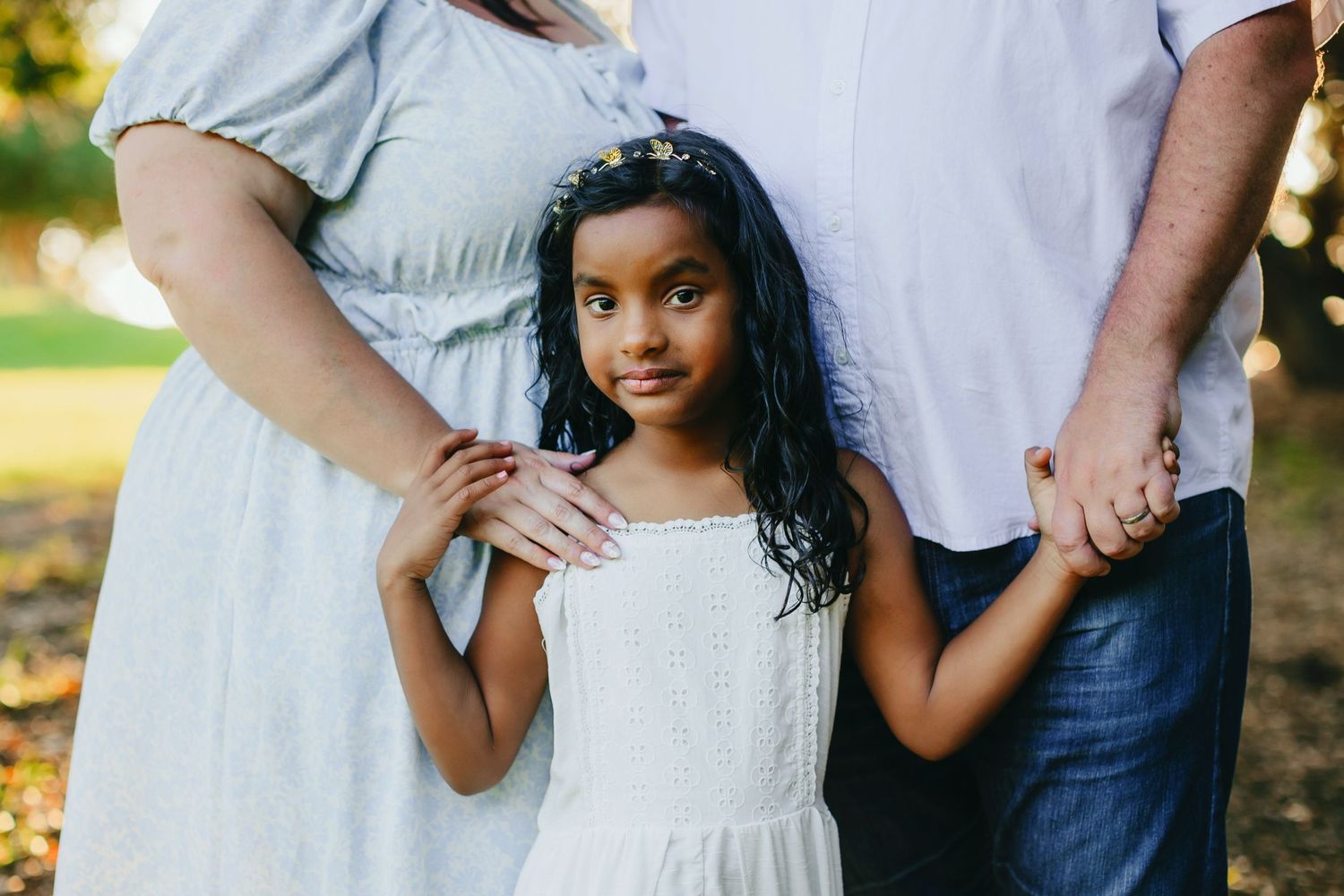 Family portrait showing parents holding hands with child in white dress outdoors.