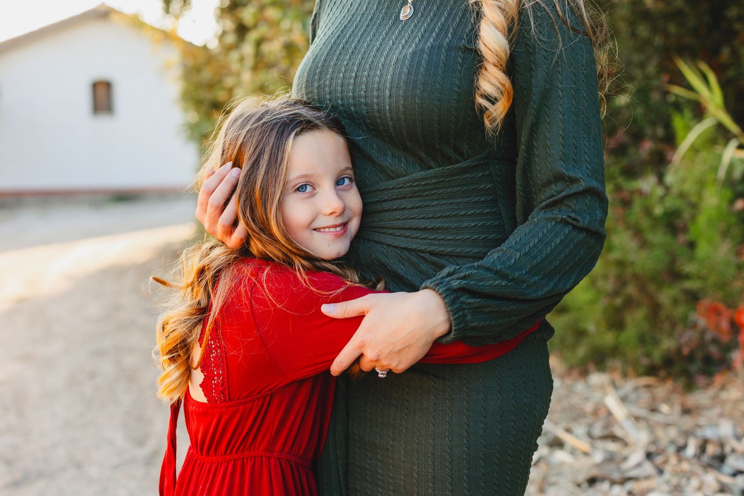 Child in red sweater hugging someone wearing a green knit dress outdoors.